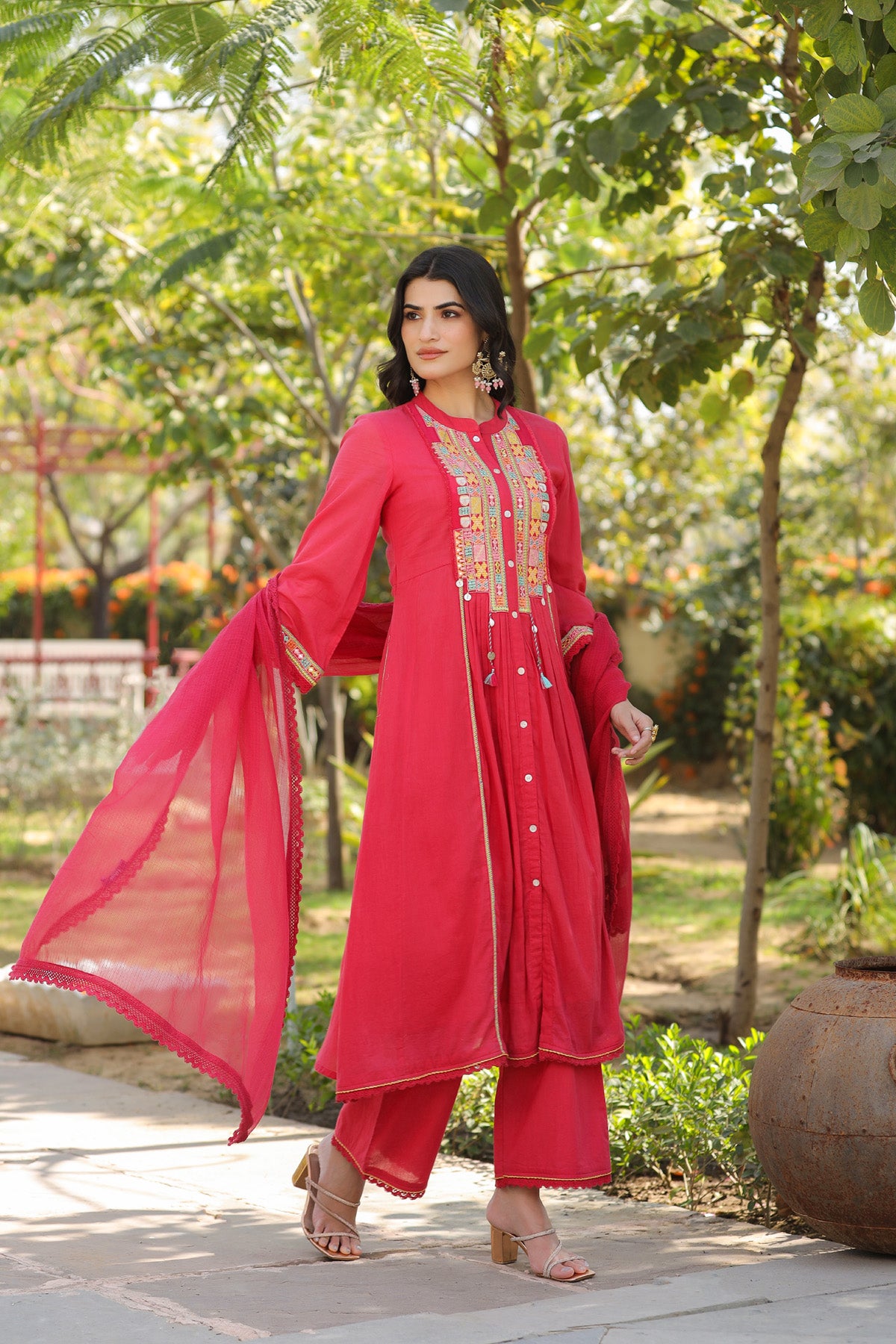 Woman in a red traditional outfit standing outdoors with greenery in the background
