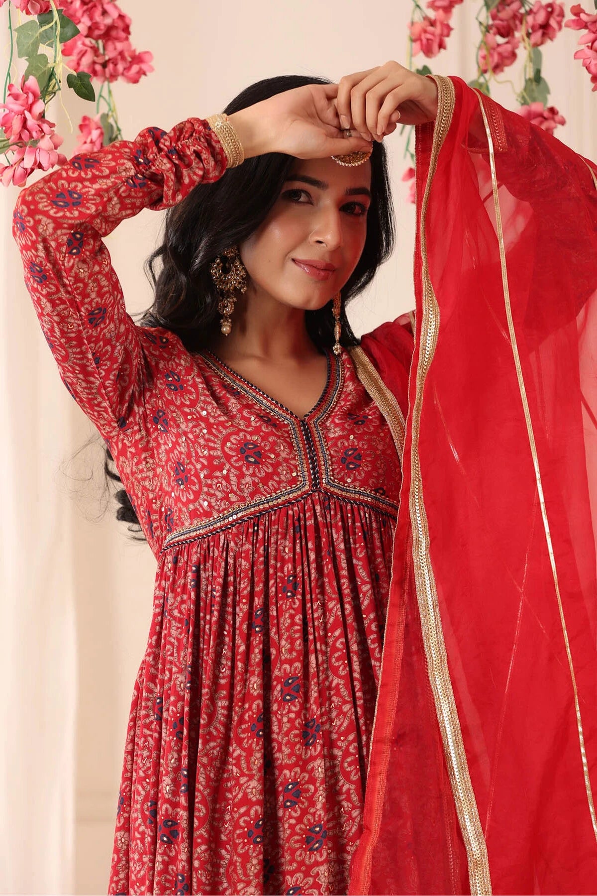 Woman in a red patterned dress holding a red dupatta against a floral background