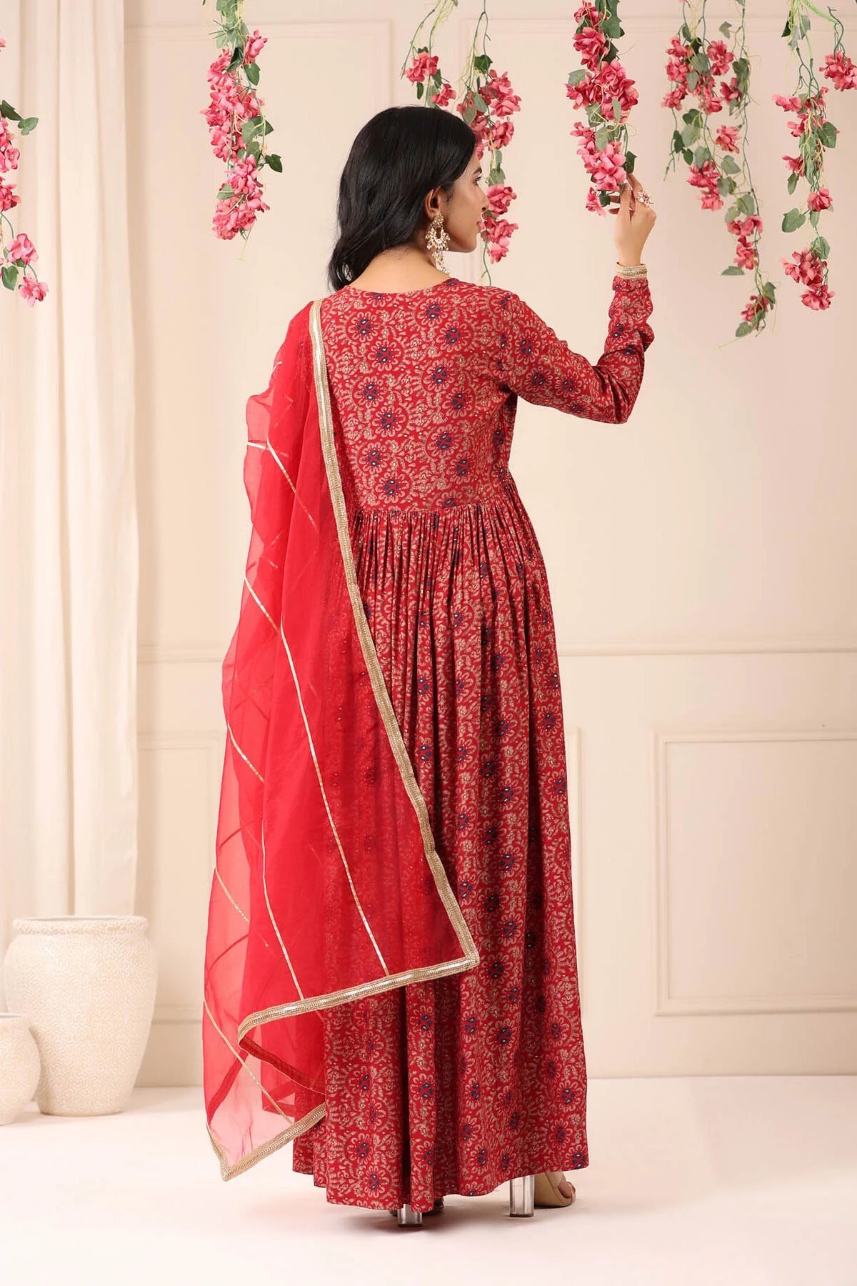Woman in a red and black patterned dress with a matching dupatta, standing against a light-colored wall with floral decorations.