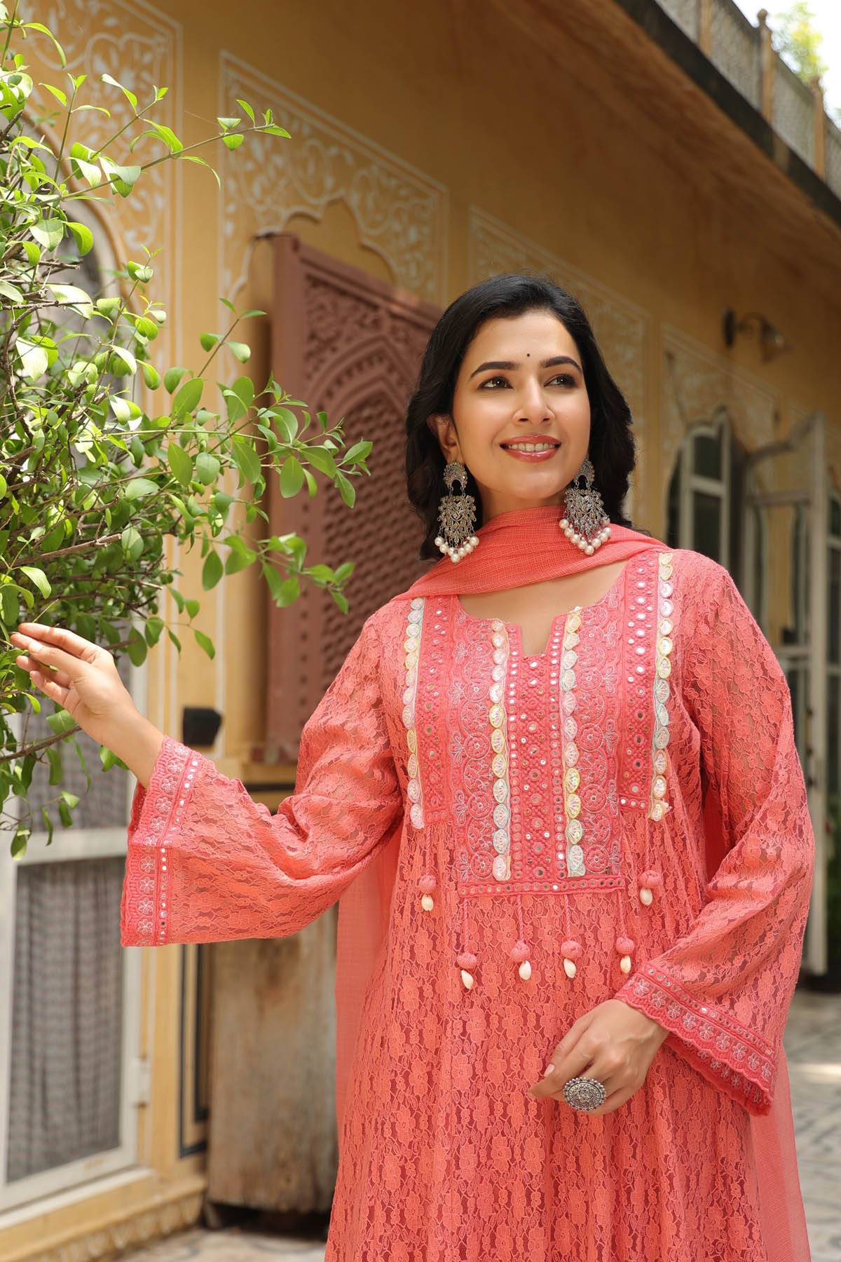 Woman in a coral traditional outfit standing outdoors with a building and tree in the background