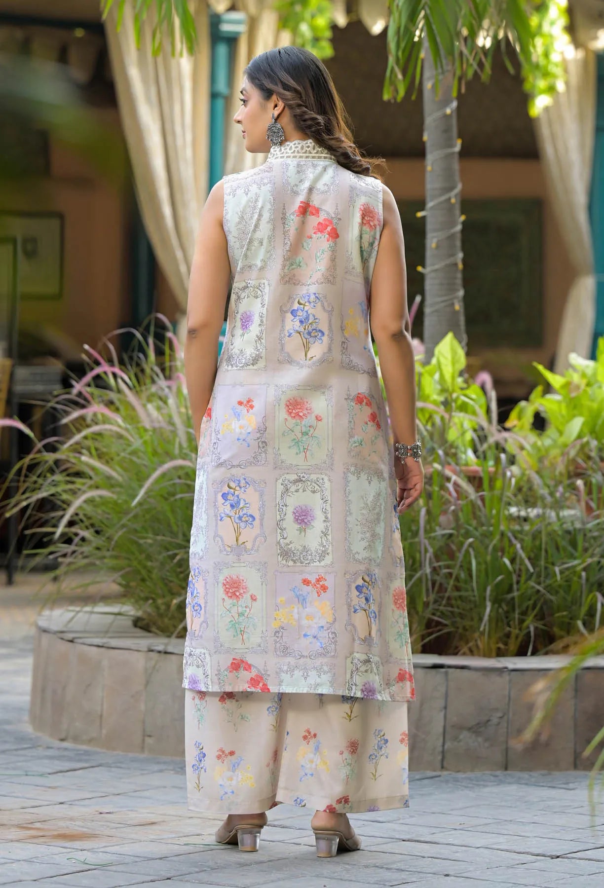 Woman wearing a floral embroidered outfit standing outdoors with plants and a building in the background.