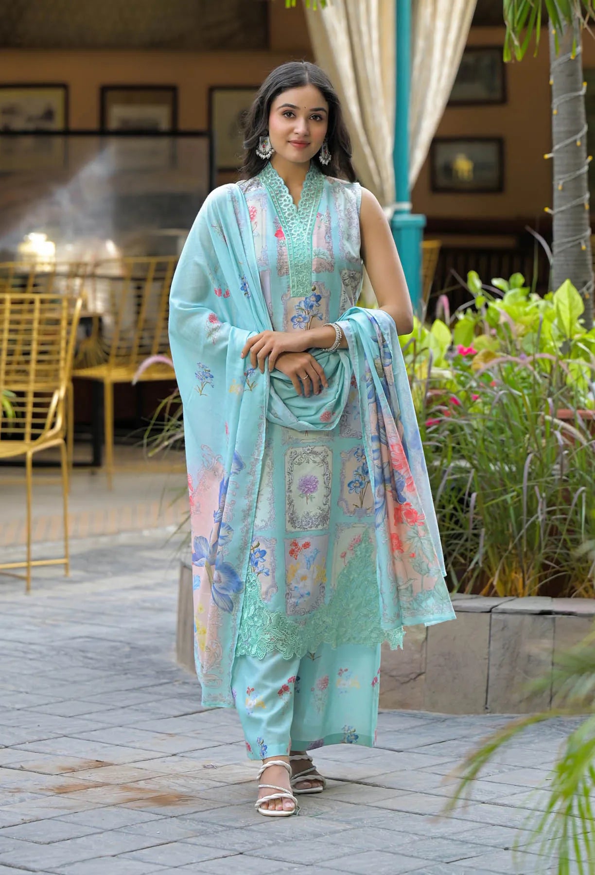 Woman in a light blue embroidered outfit standing outdoors with plants and furniture in the background