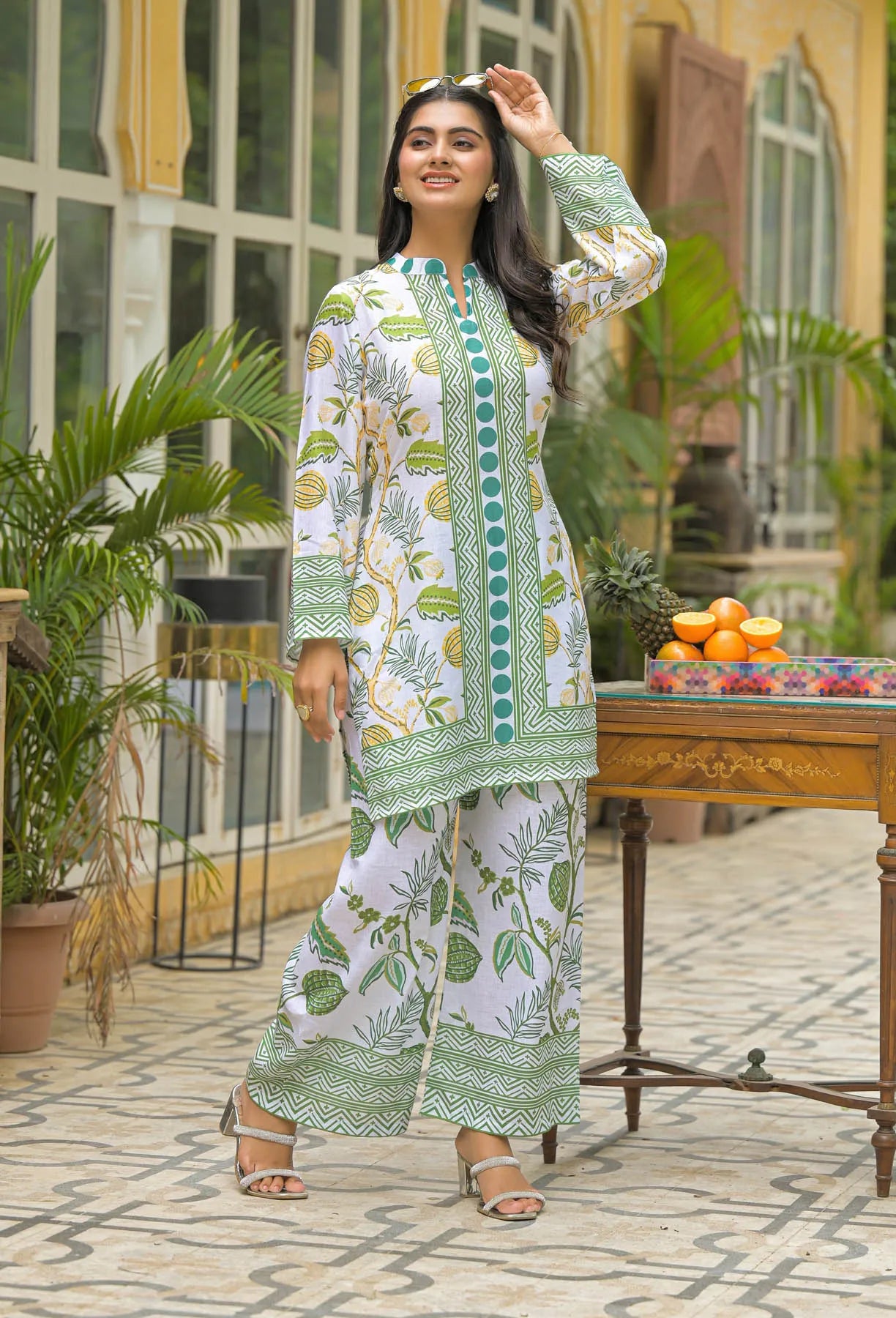 Woman in a green and white patterned outfit standing outdoors with plants and a table in the background.
