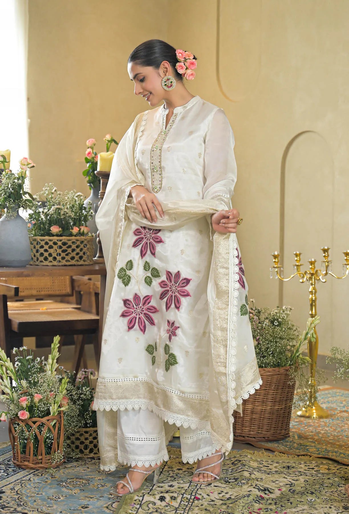 Woman in a white embroidered traditional outfit standing in a decorated room with flowers and candles.