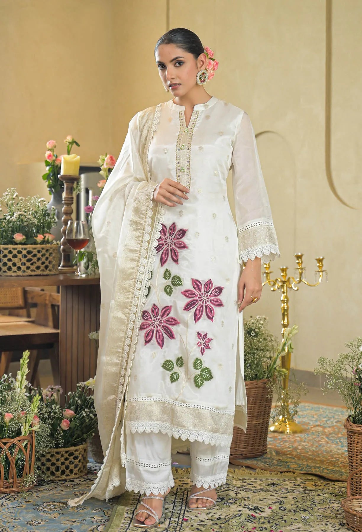 Woman in a white embroidered outfit standing in an elegant indoor setting with floral decorations.
