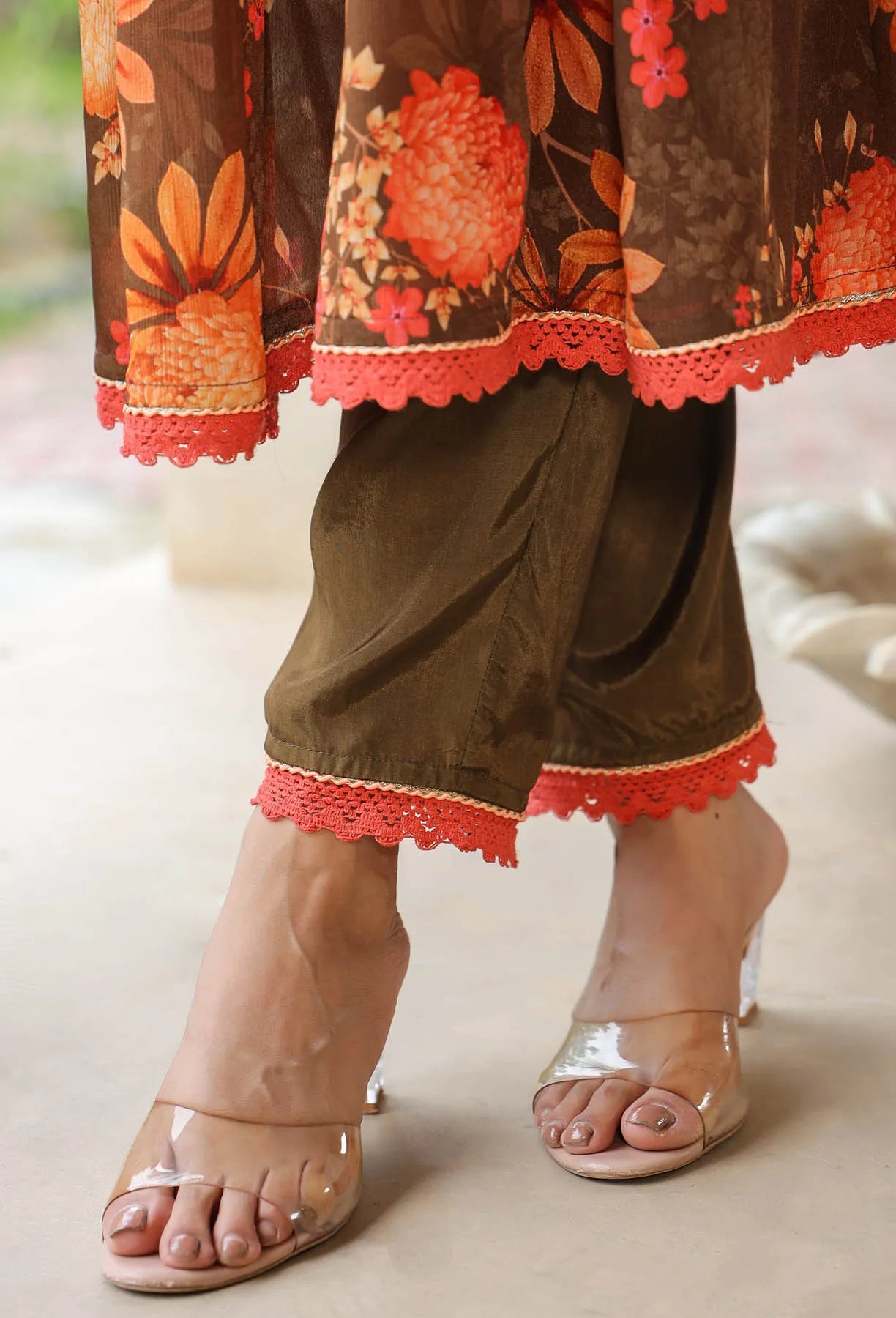 Traditional outfit with floral top and brown pants with red lace trim, paired with clear high-heeled sandals.