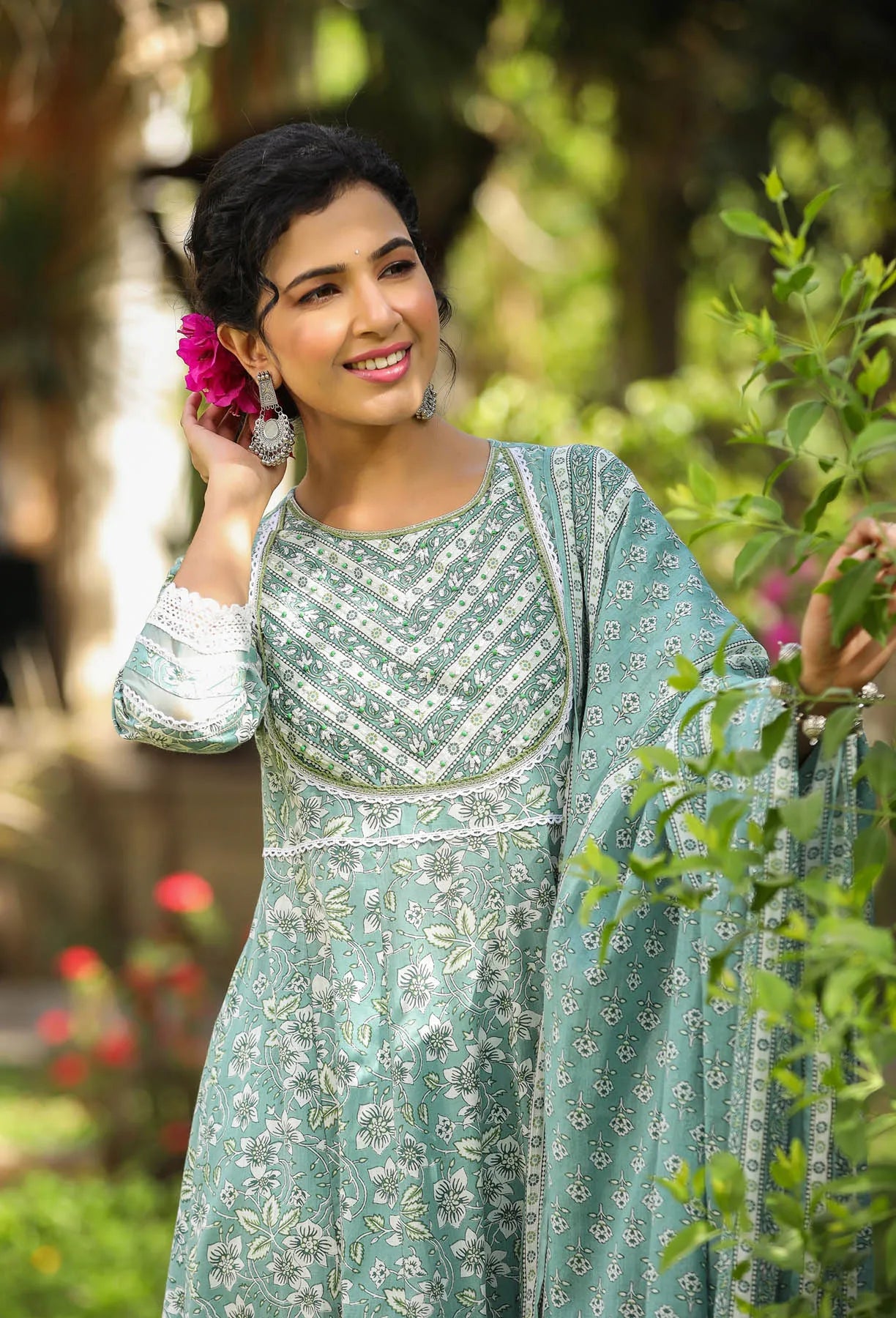 Woman in a light blue traditional outfit with floral patterns, standing outdoors with greenery.