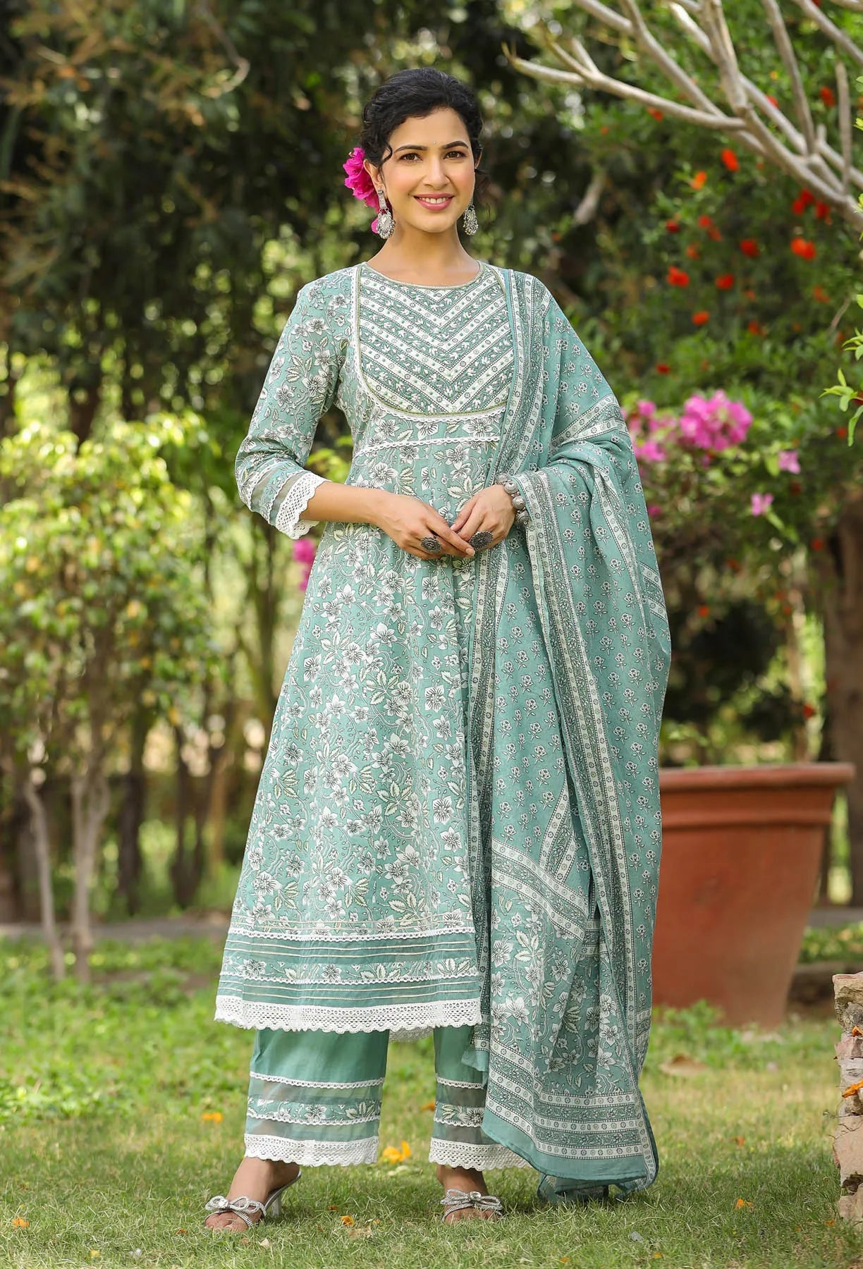 Woman in a traditional green outfit standing outdoors with trees and flowers in the background