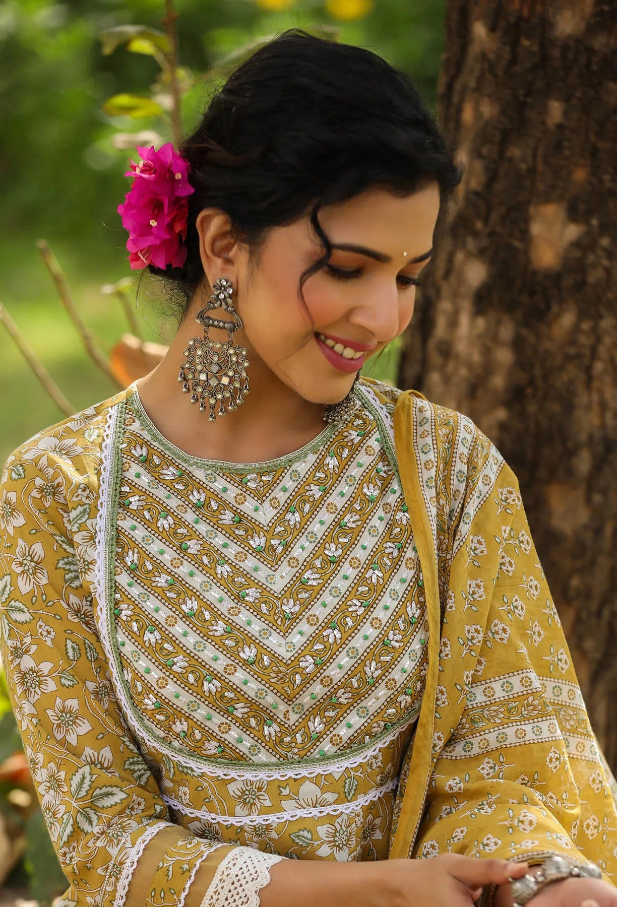 Woman in traditional yellow and white embroidered outfit with floral hair accessory, sitting outdoors.