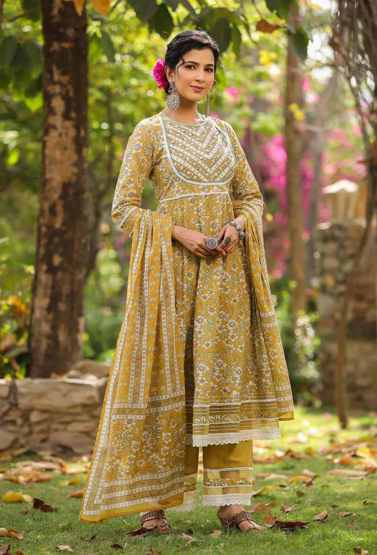 Woman in a yellow traditional outfit standing outdoors with greenery in the background