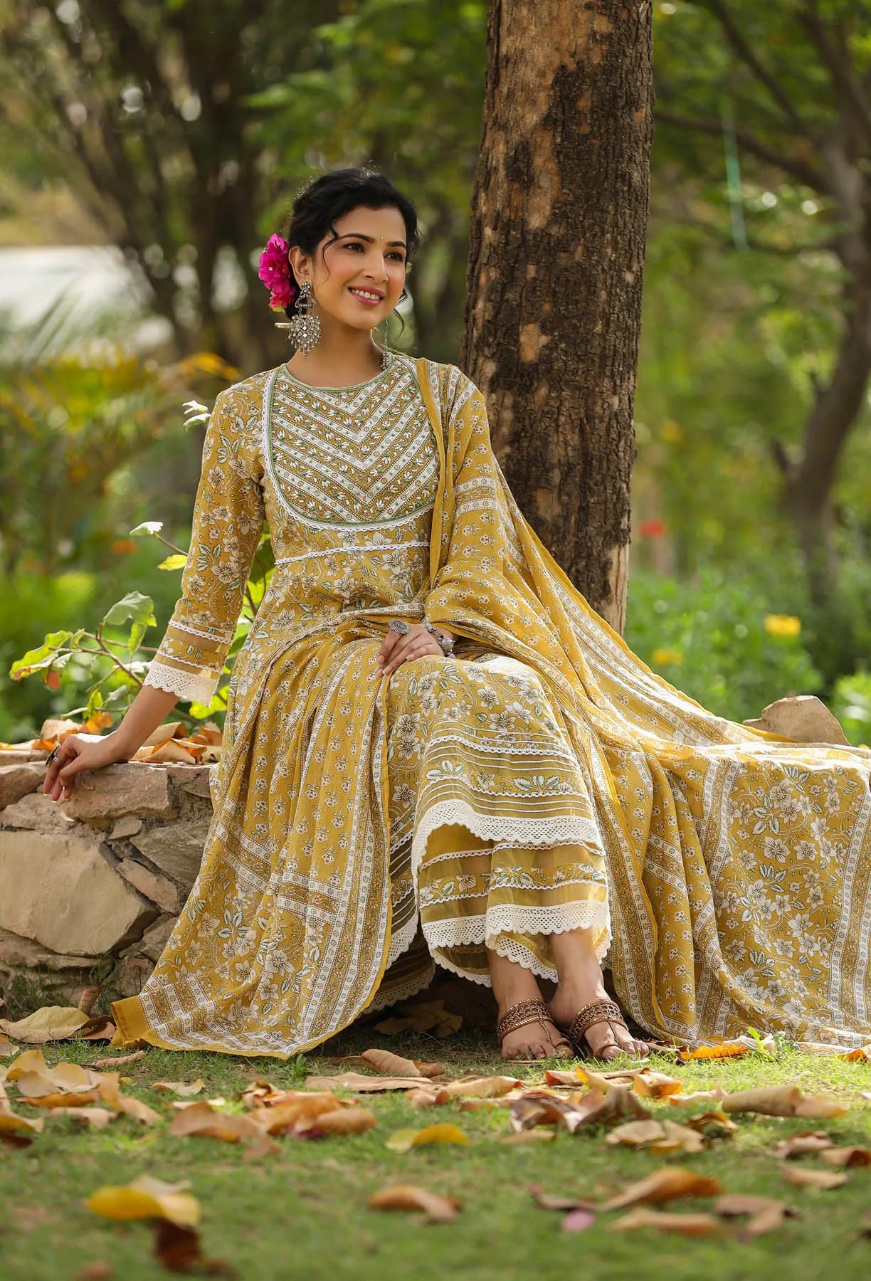 Woman in a yellow and white traditional outfit sitting under a tree in a garden.