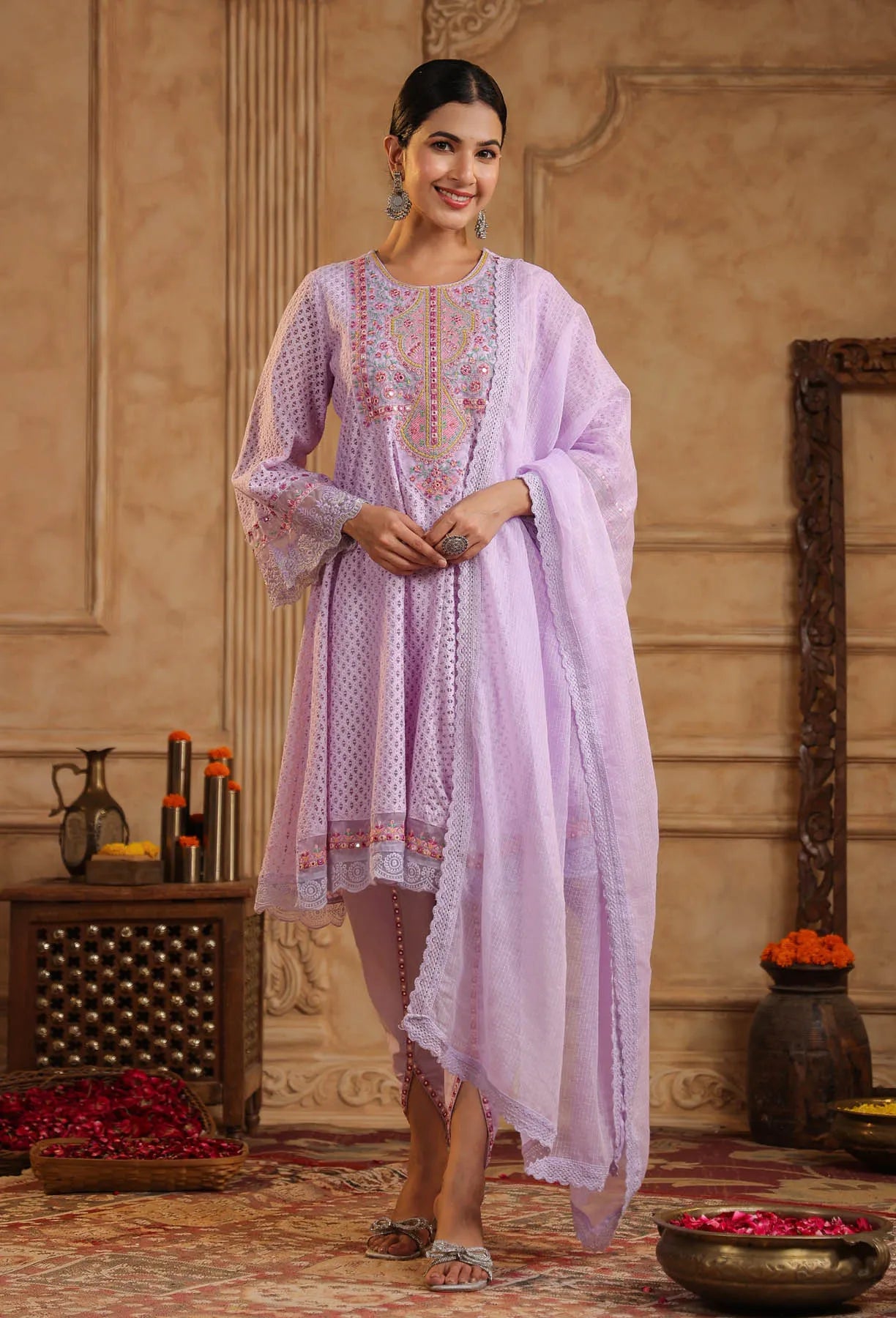 Woman in a light purple traditional outfit with intricate patterns standing in a decorated room.