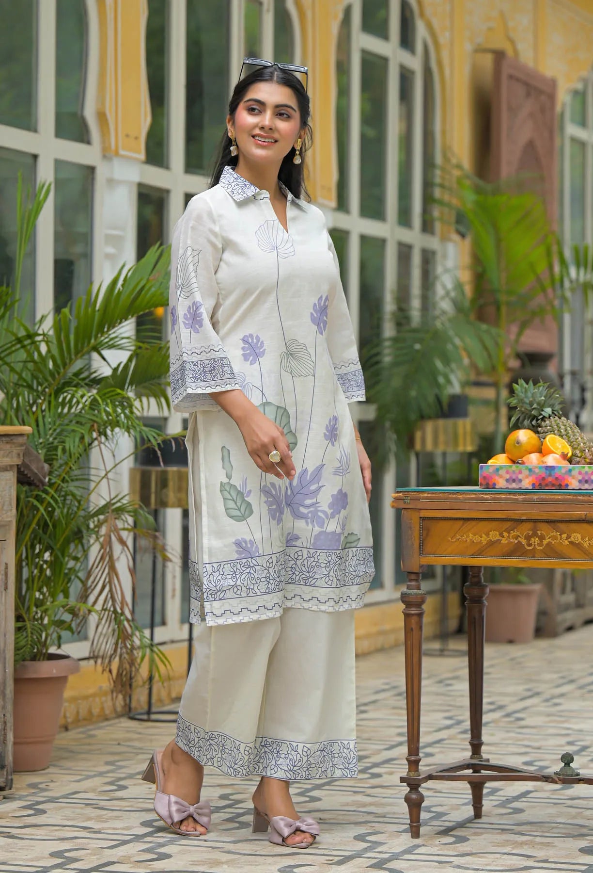 Woman in a white floral outfit standing in front of a building with plants and a table.