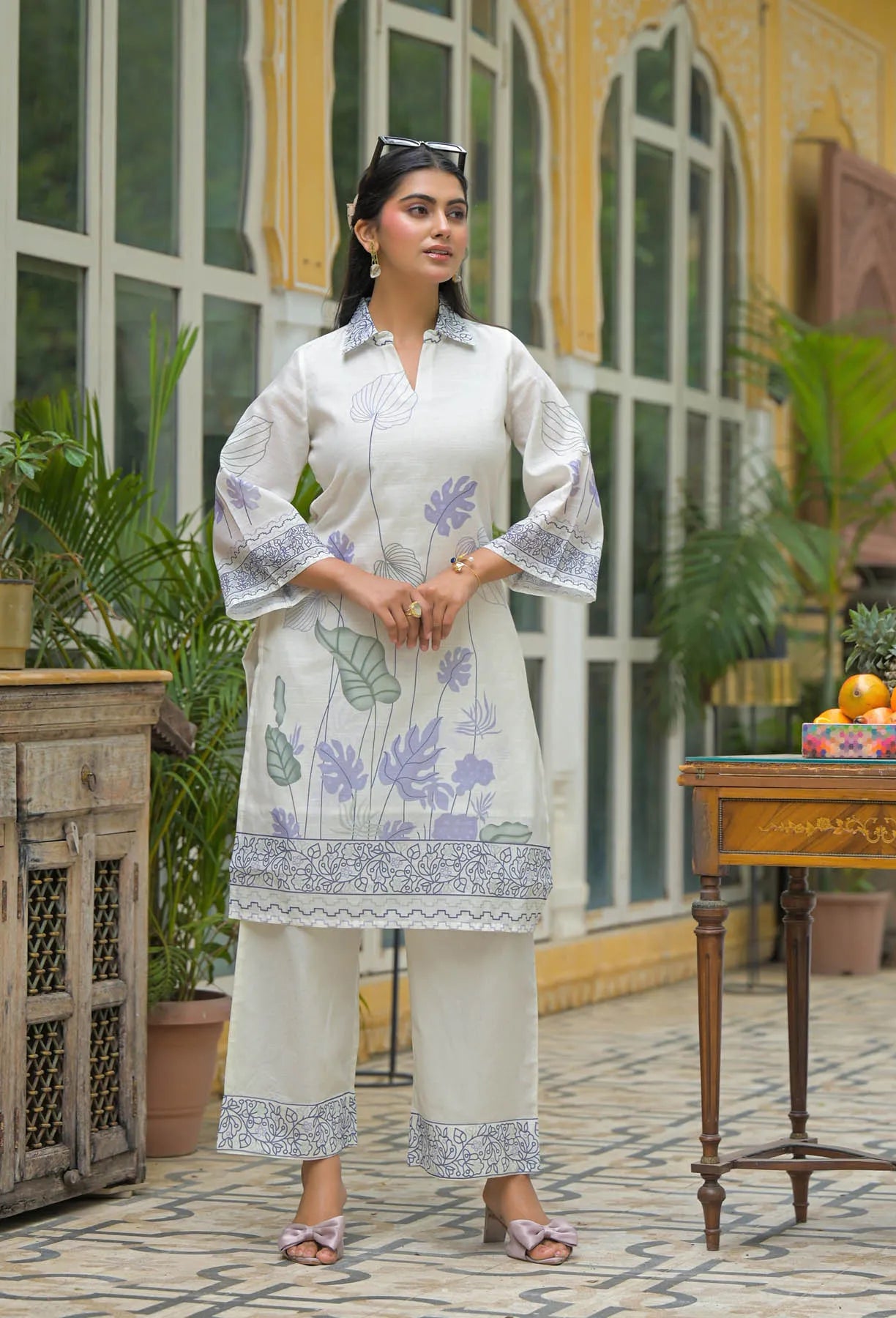 Woman wearing a white embroidered outfit standing in an outdoor setting with plants and a table.