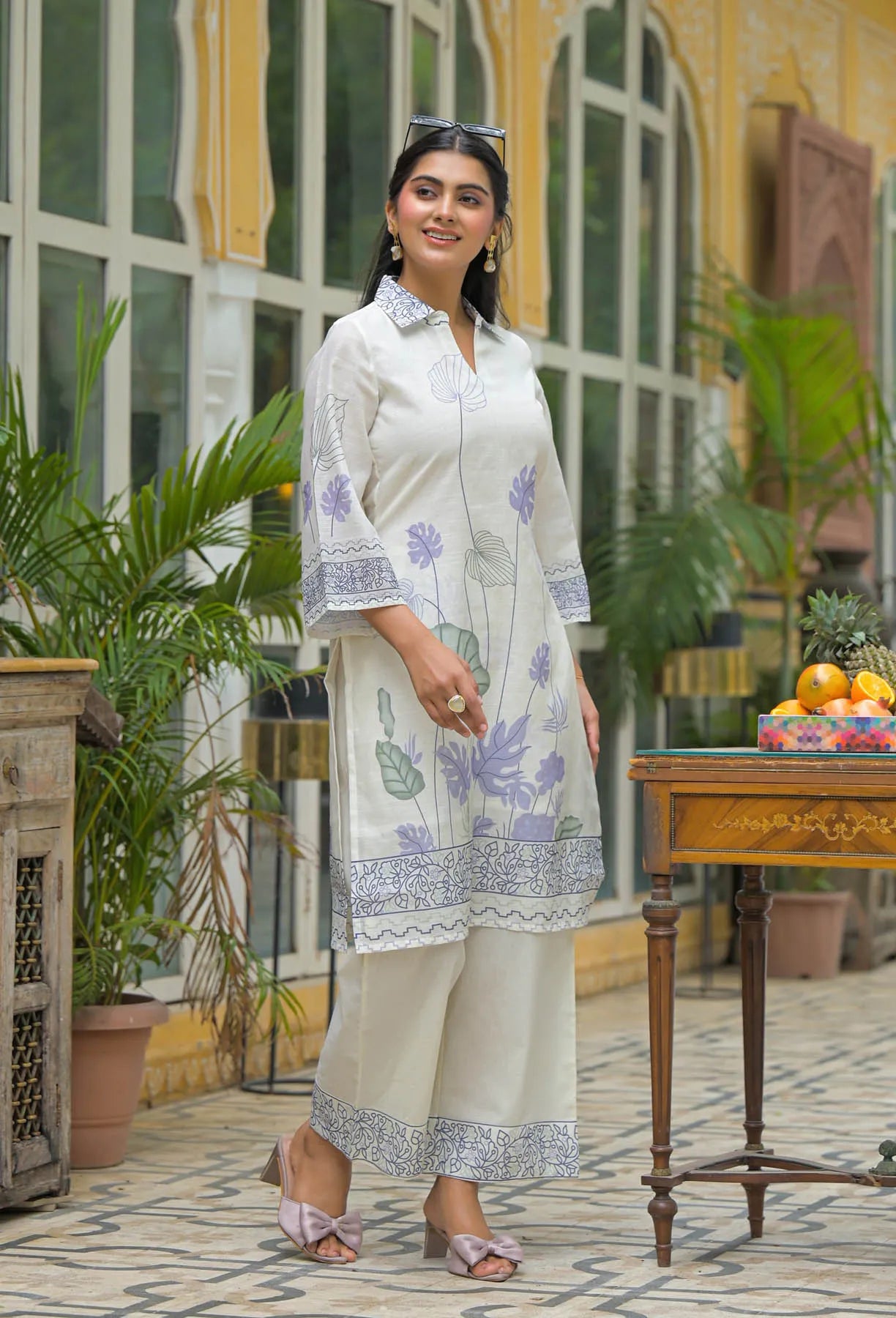 Woman in a white traditional outfit with floral patterns standing in a decorative outdoor setting.
