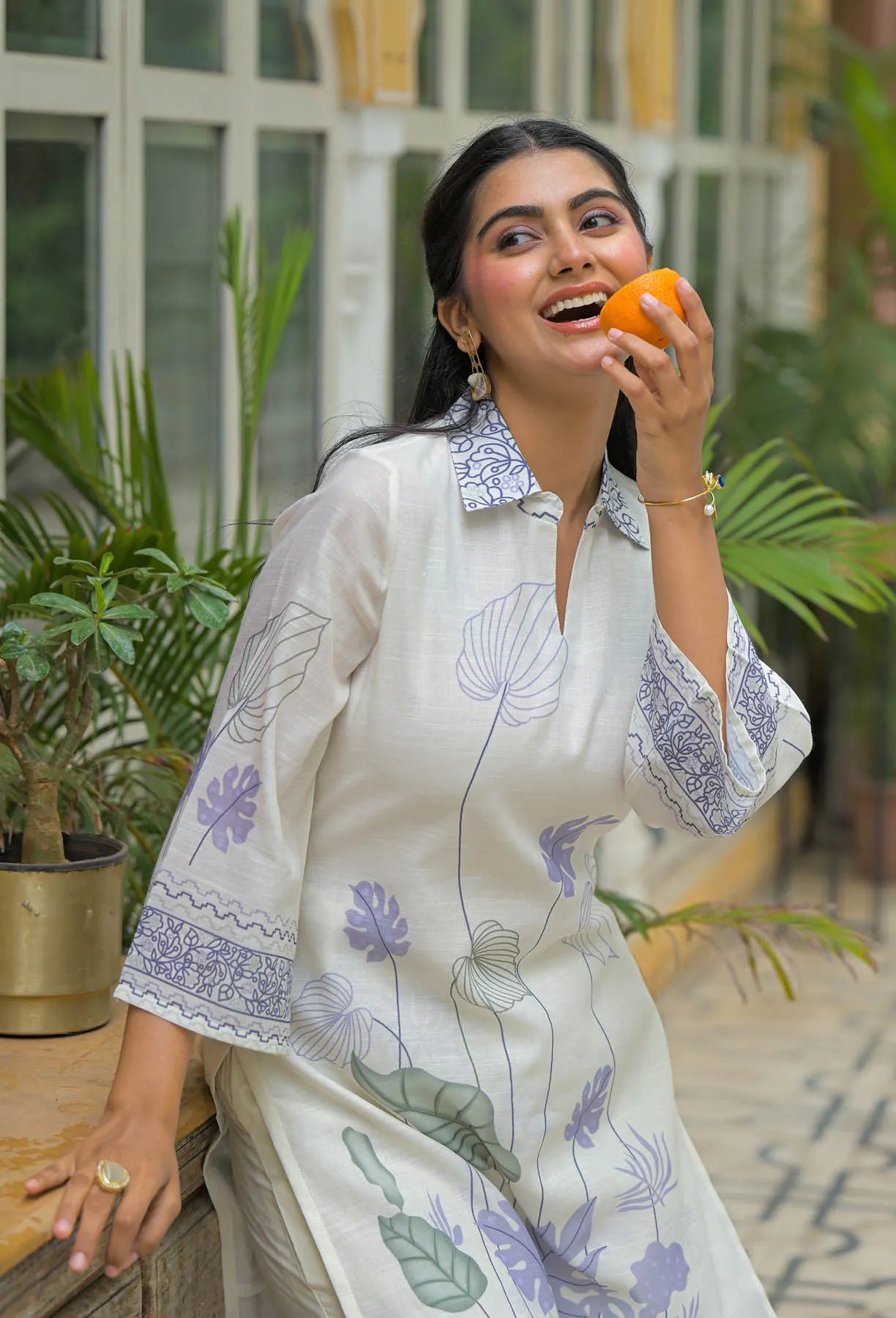 Woman in a white dress with floral patterns holding an orange outdoors.