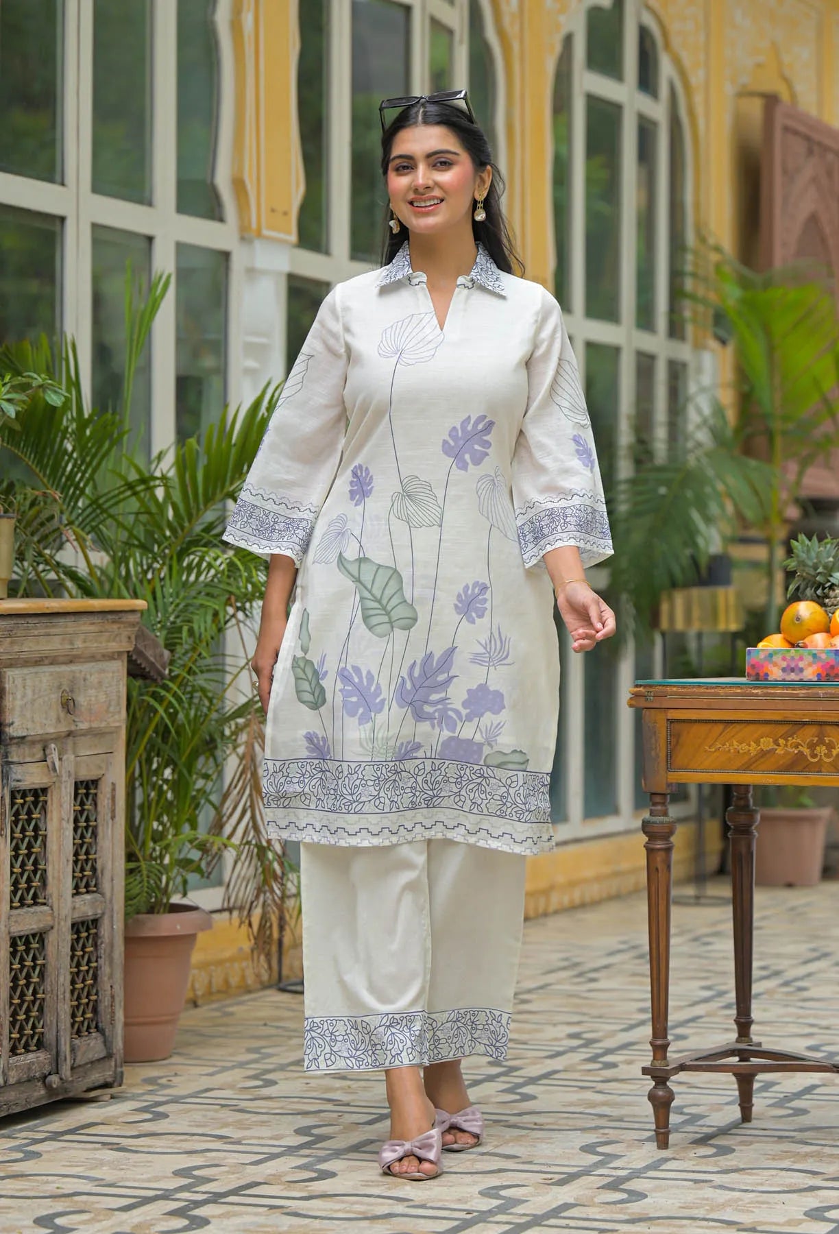 Woman in a white floral outfit standing in an outdoor setting with plants and a table.