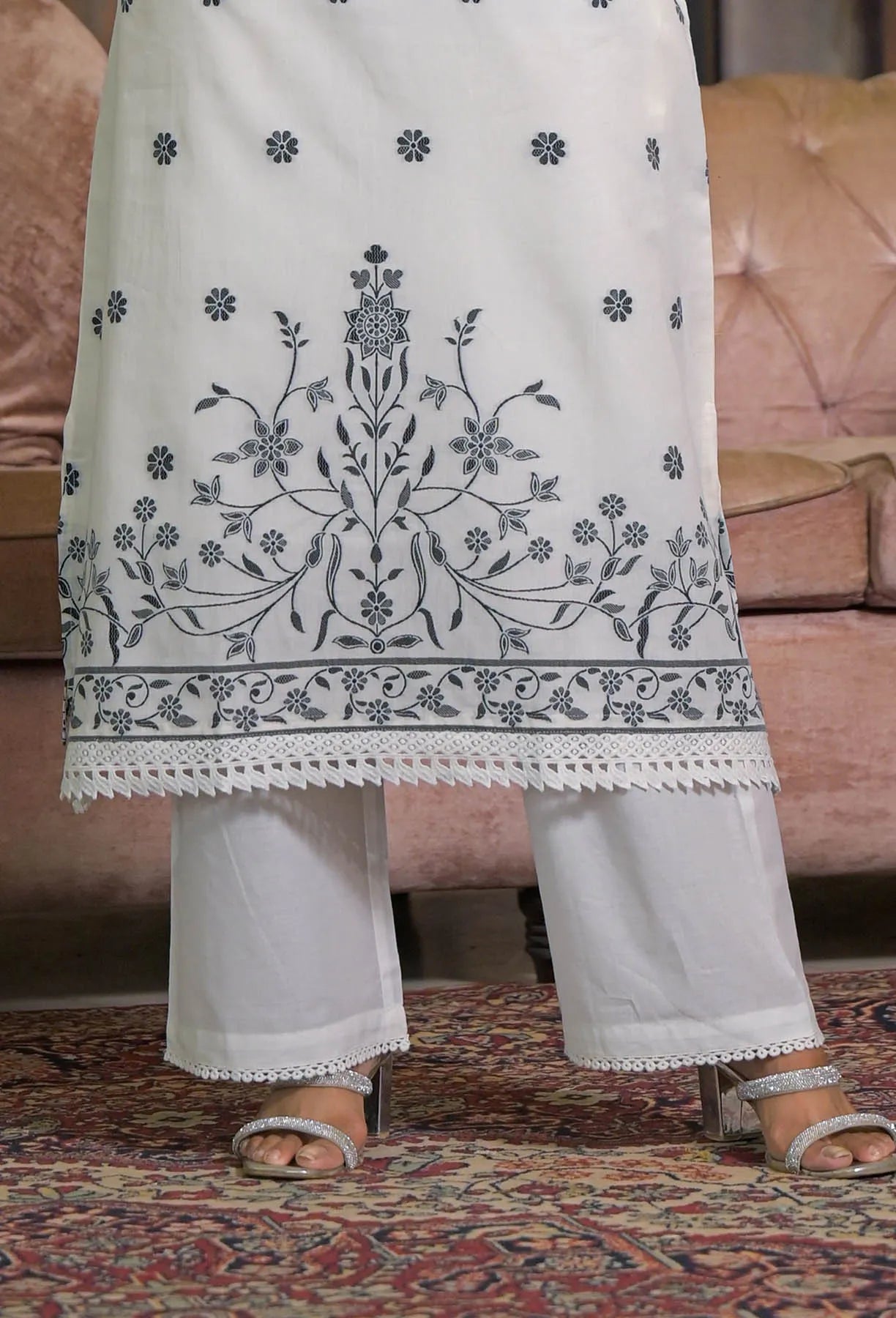 White embroidered kurta with black patterns worn by a person, sitting on a patterned rug.