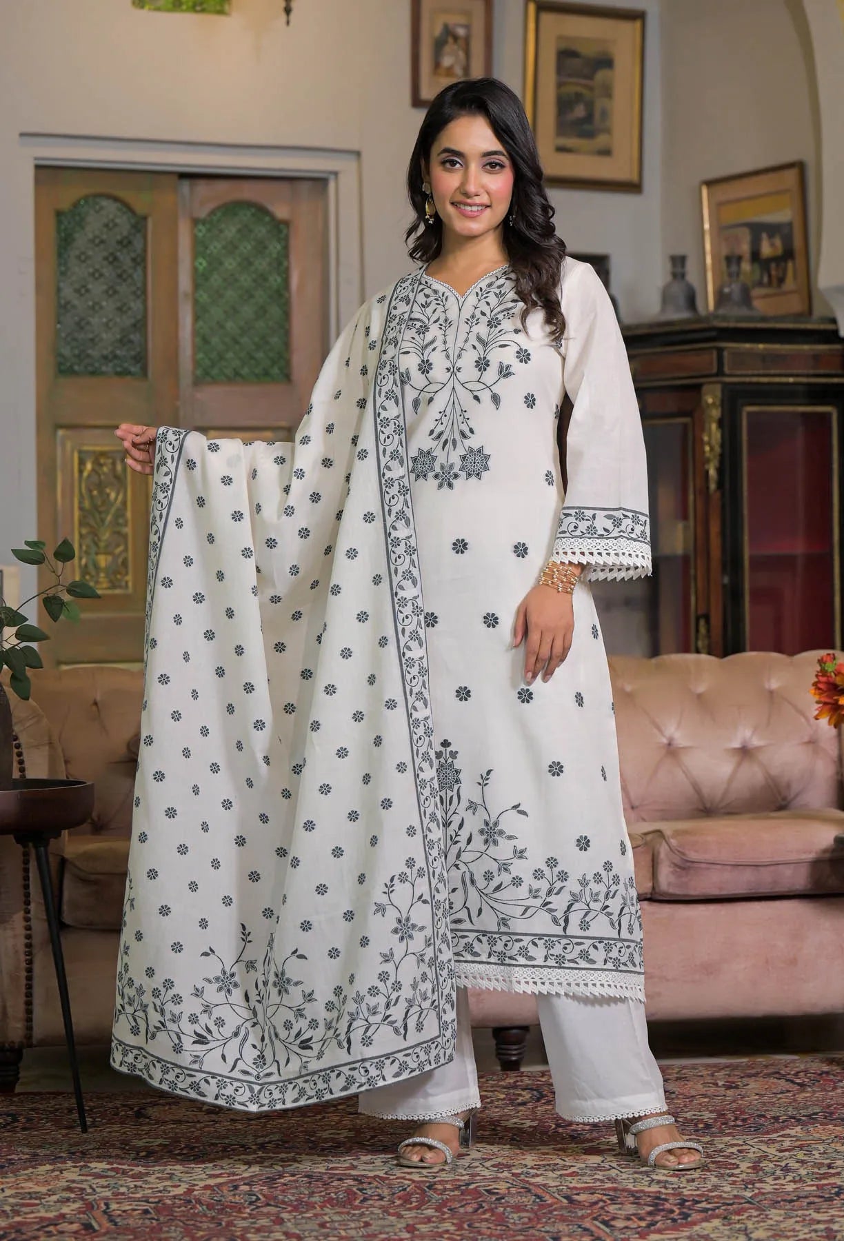 Woman in a white and gray embroidered traditional outfit standing in a decorated room.