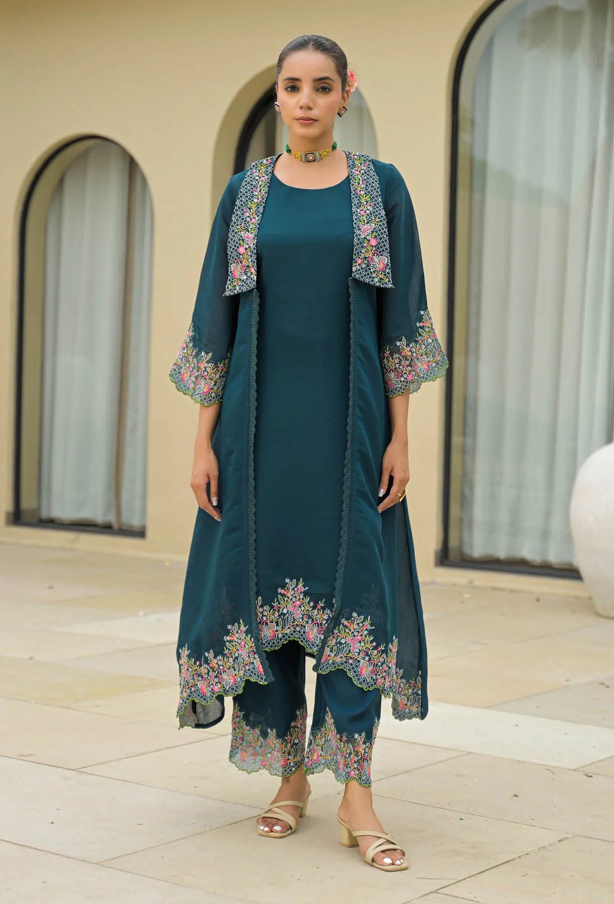 Woman wearing a teal embroidered outfit standing in front of a building with large windows.