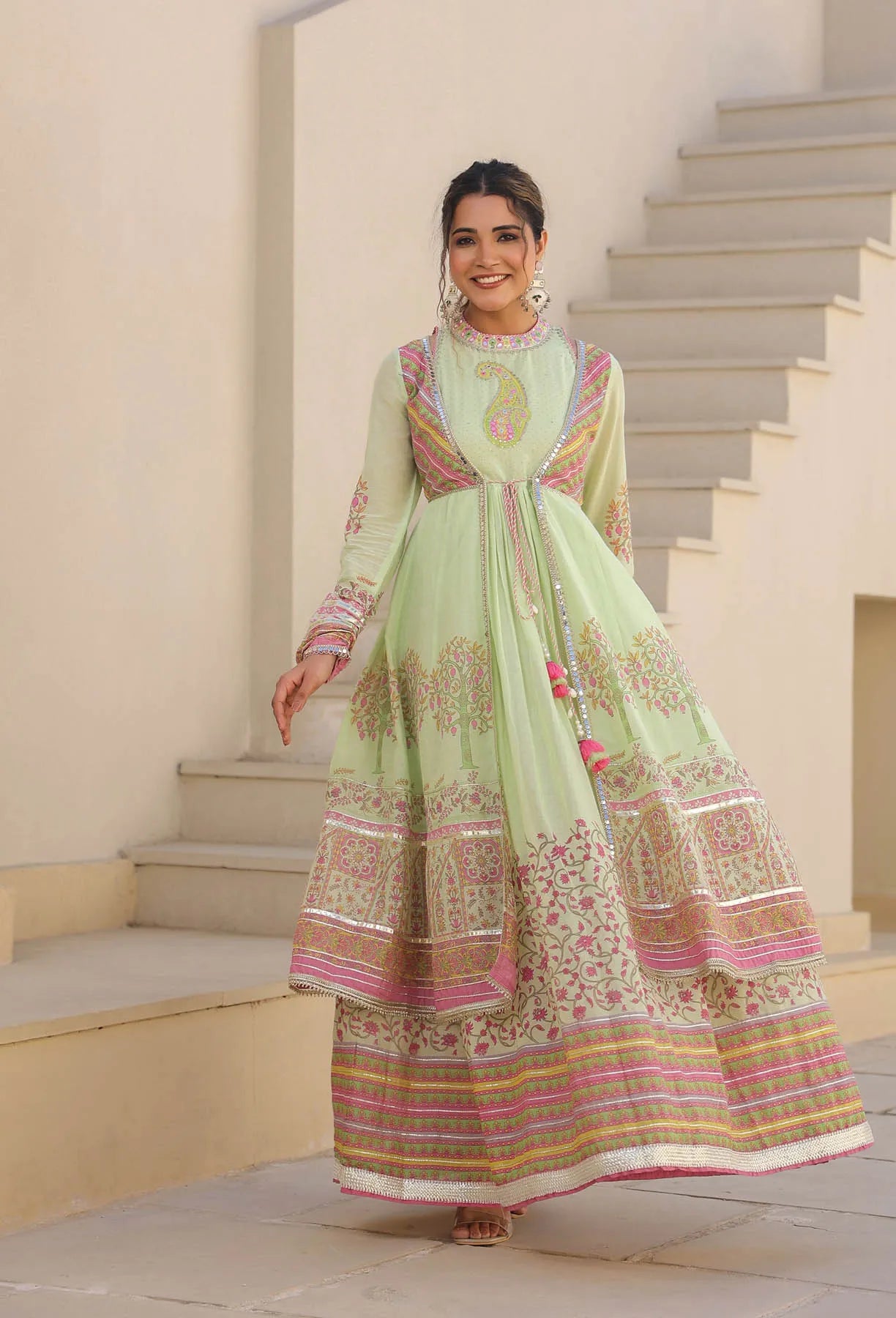 Woman in a light green traditional outfit with pink and gold patterns standing on steps.