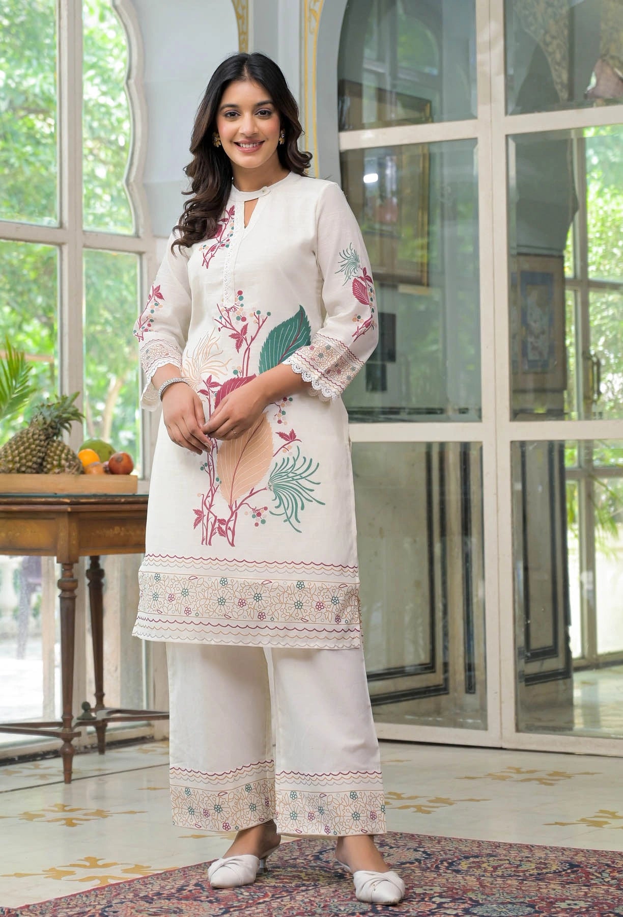 Woman wearing a white embroidered outfit standing in a room with large windows.