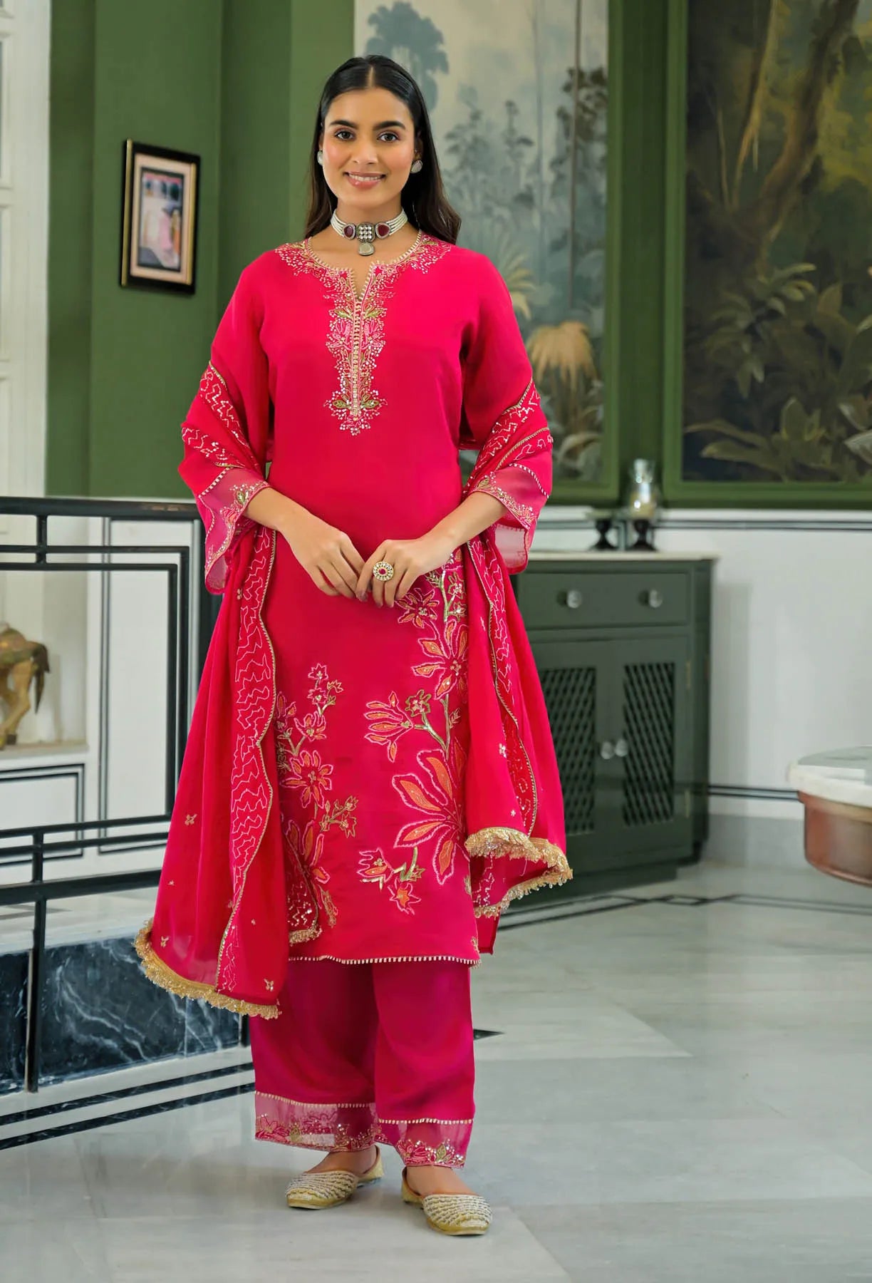 Woman in a bright pink traditional outfit with floral patterns in a room with green walls and decorative items.
