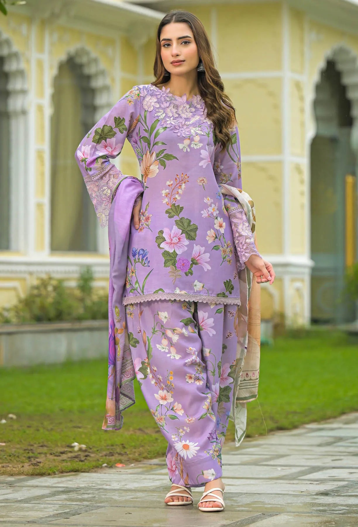 Woman in a floral purple outfit standing in front of a building with greenery.
