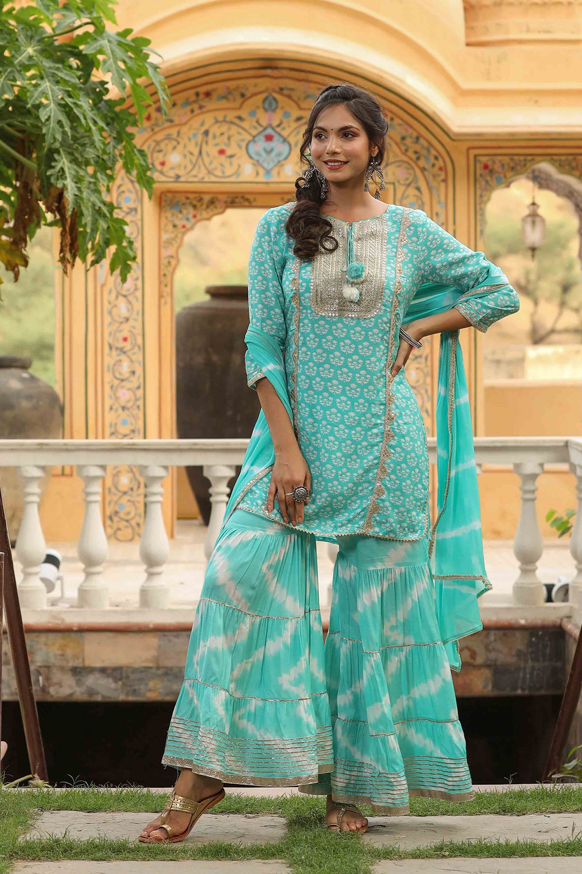 Woman in a turquoise traditional outfit standing outdoors with decorative architecture in the background