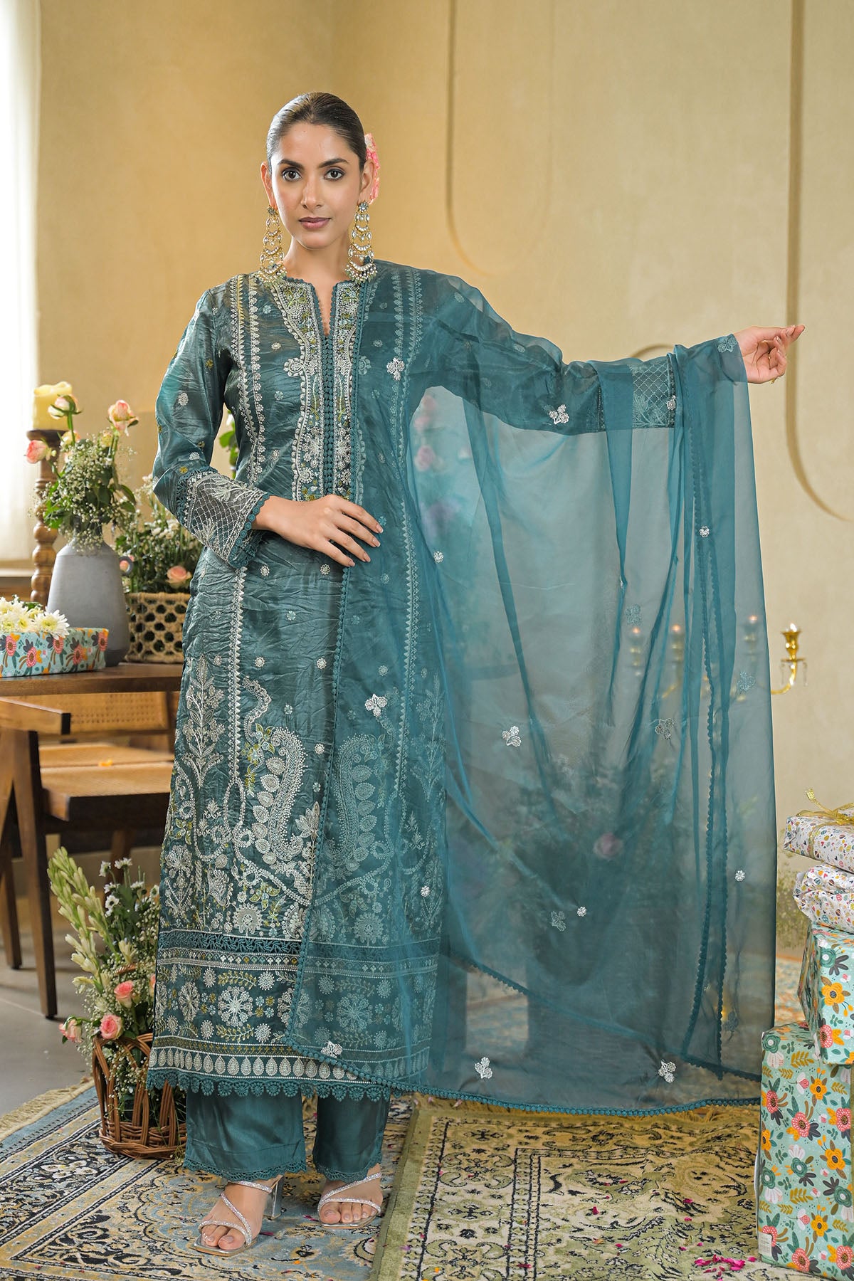 Woman in a teal embroidered traditional outfit with a matching dupatta in a decorated room.