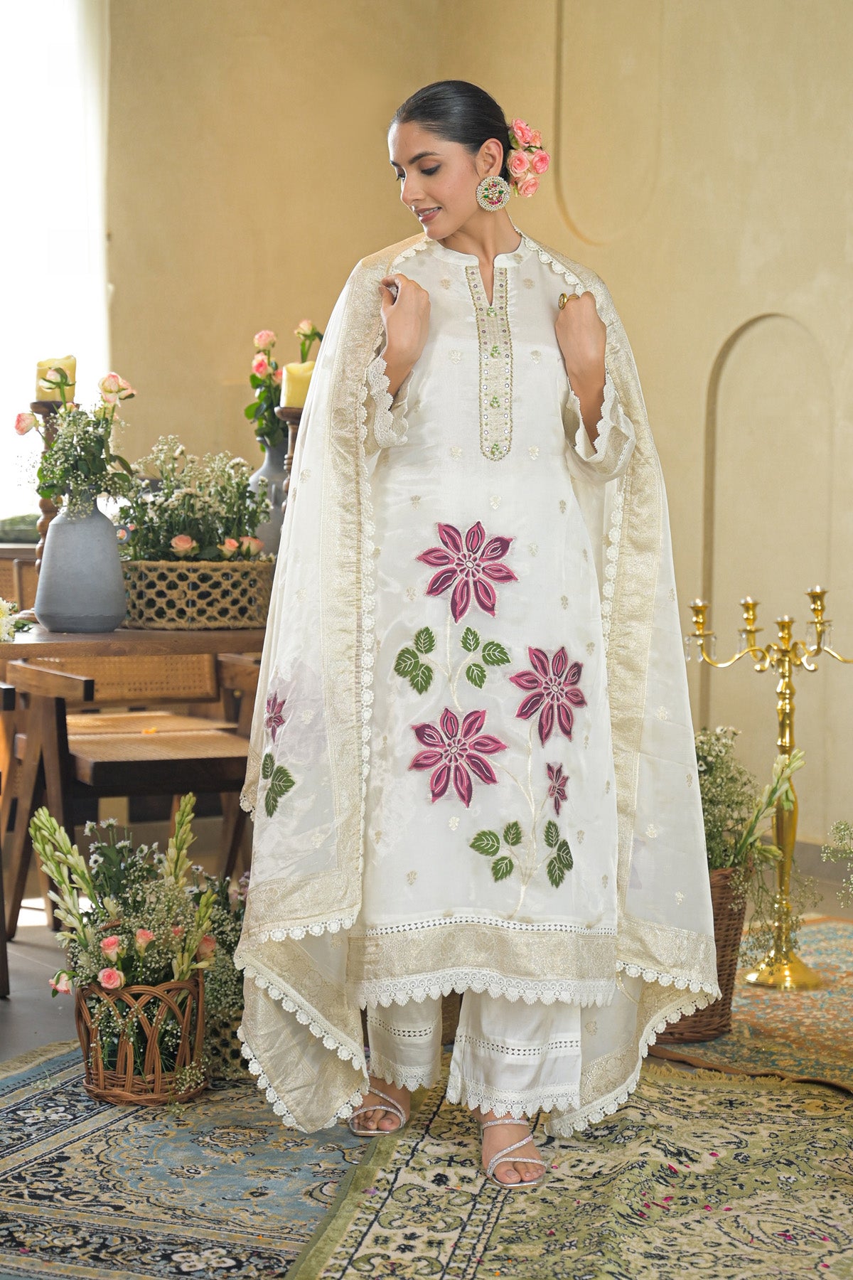 Woman in a white embroidered outfit standing in an elegant room with floral decorations.