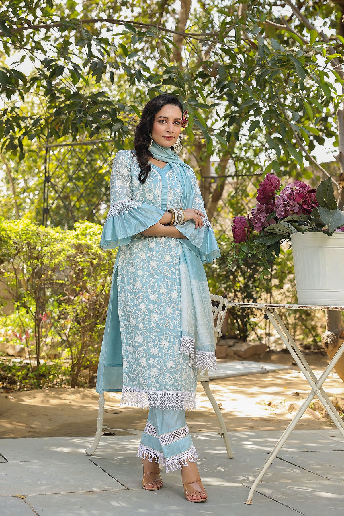 Woman in a light blue traditional outfit standing outdoors with greenery and flowers.