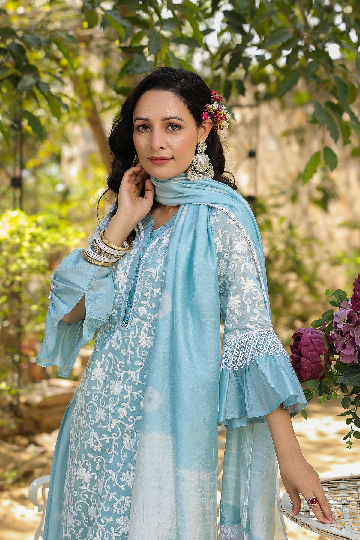 Woman in a light blue traditional outfit with white embroidery, standing outdoors with greenery in the background.