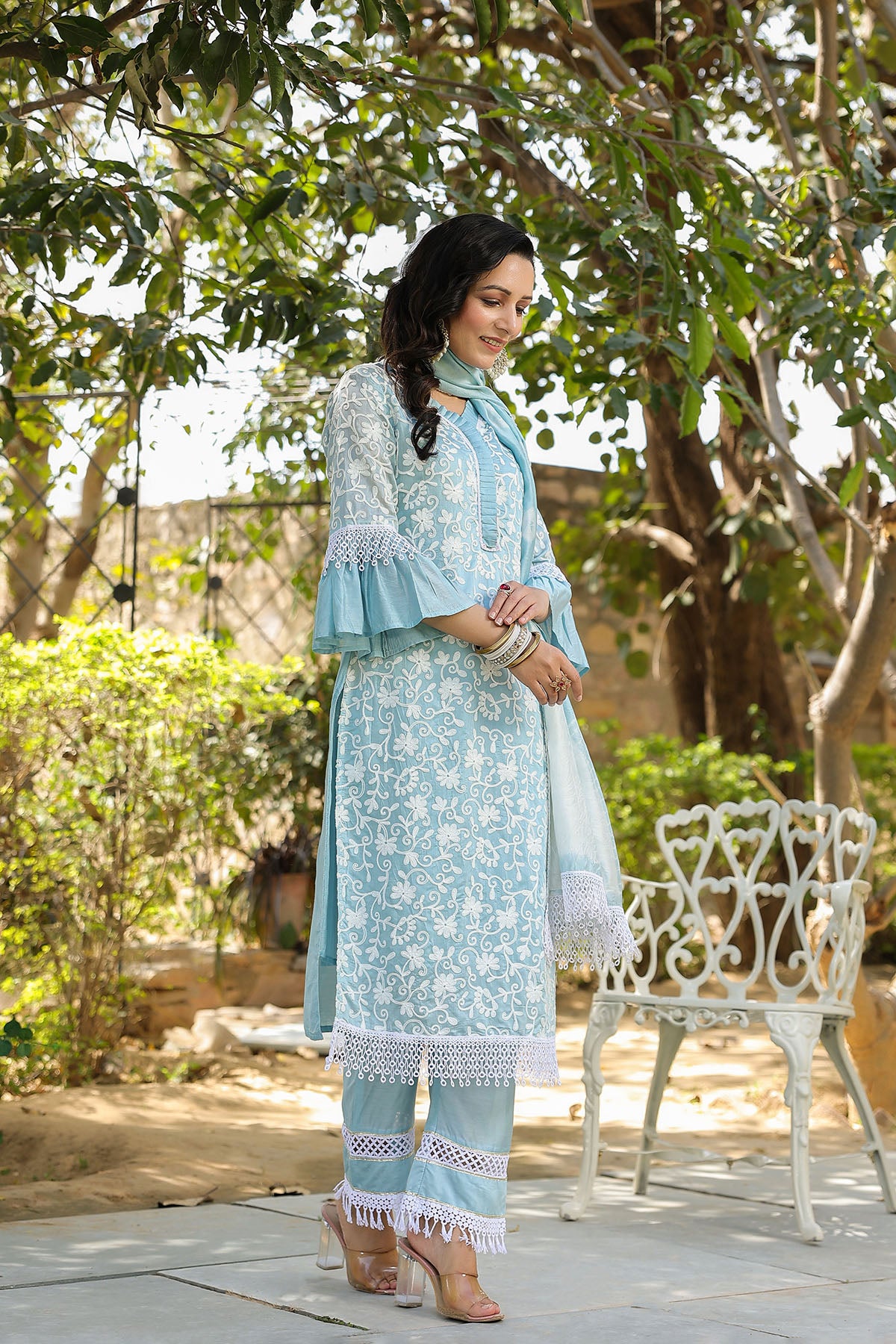 Woman in a light blue traditional outfit standing outdoors with greenery around.