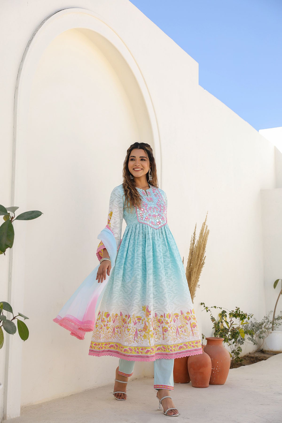 Woman in a colorful dress standing in front of a white archway with plants around.