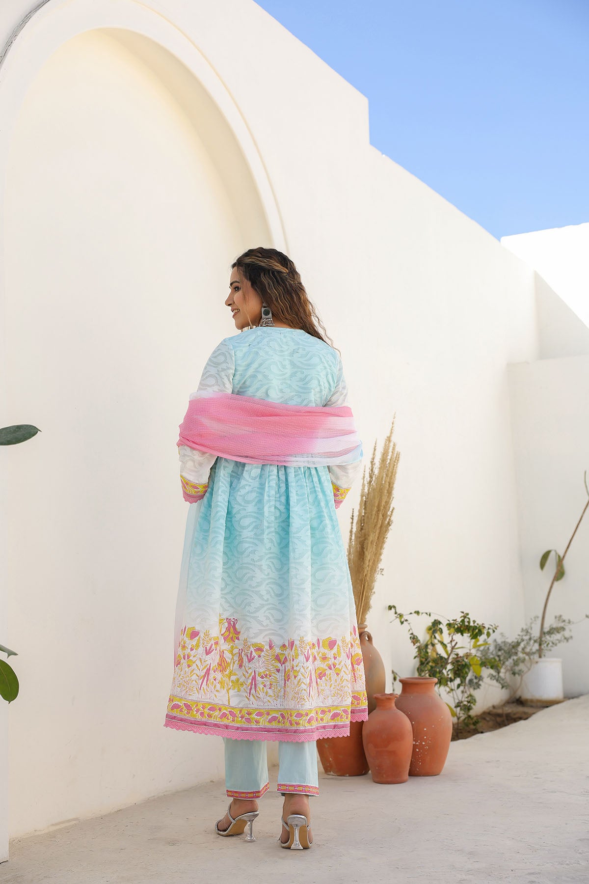 Woman in a light blue dress with floral patterns standing in a sunlit outdoor setting.