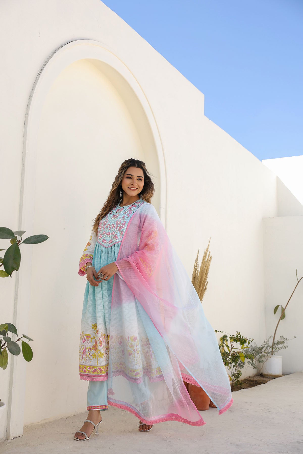 Woman in a colorful traditional outfit standing in front of a white archway with a blue sky.