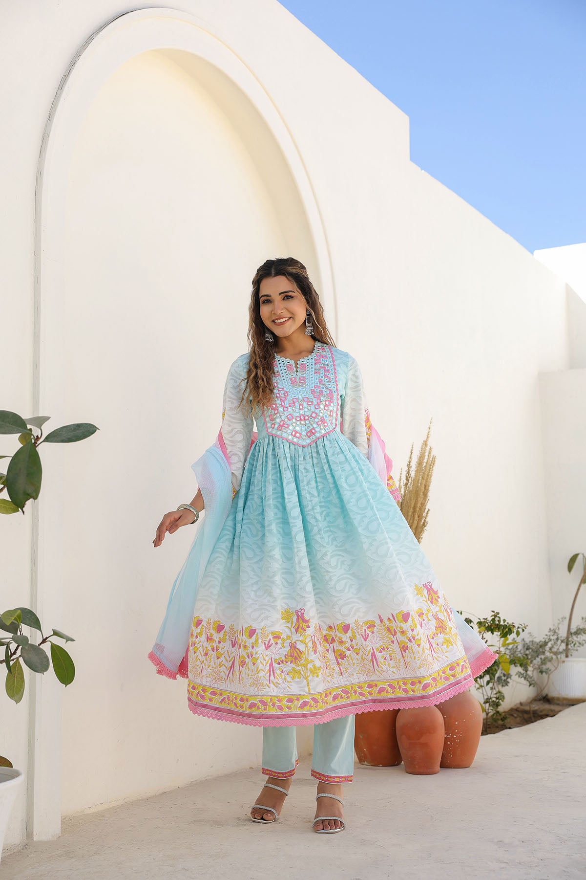 Woman in a light blue embroidered dress standing in front of a white archway with plants around.