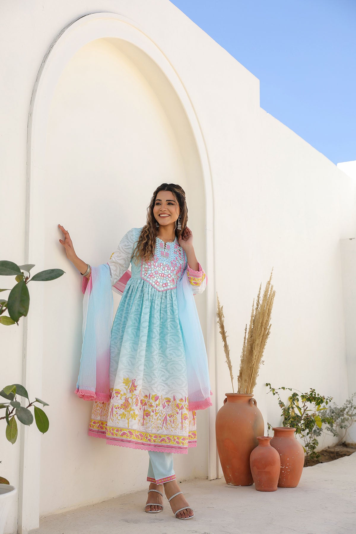 Woman in a light blue dress with floral patterns standing in front of a white archway.