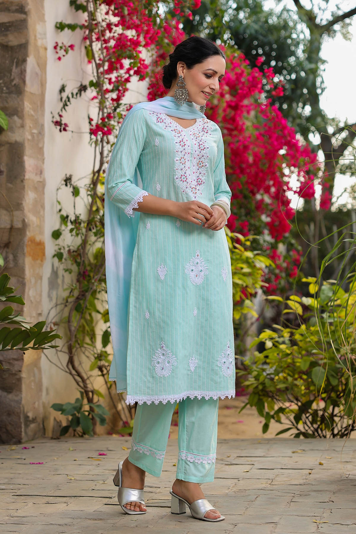 Woman wearing a light blue embroidered outfit standing in a garden with pink flowers.