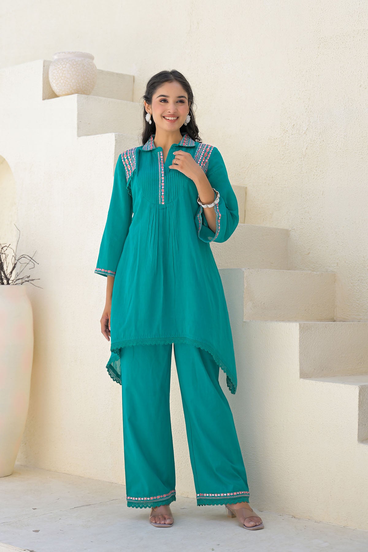 Woman wearing a teal traditional outfit with white embroidery in a minimalistic setting.