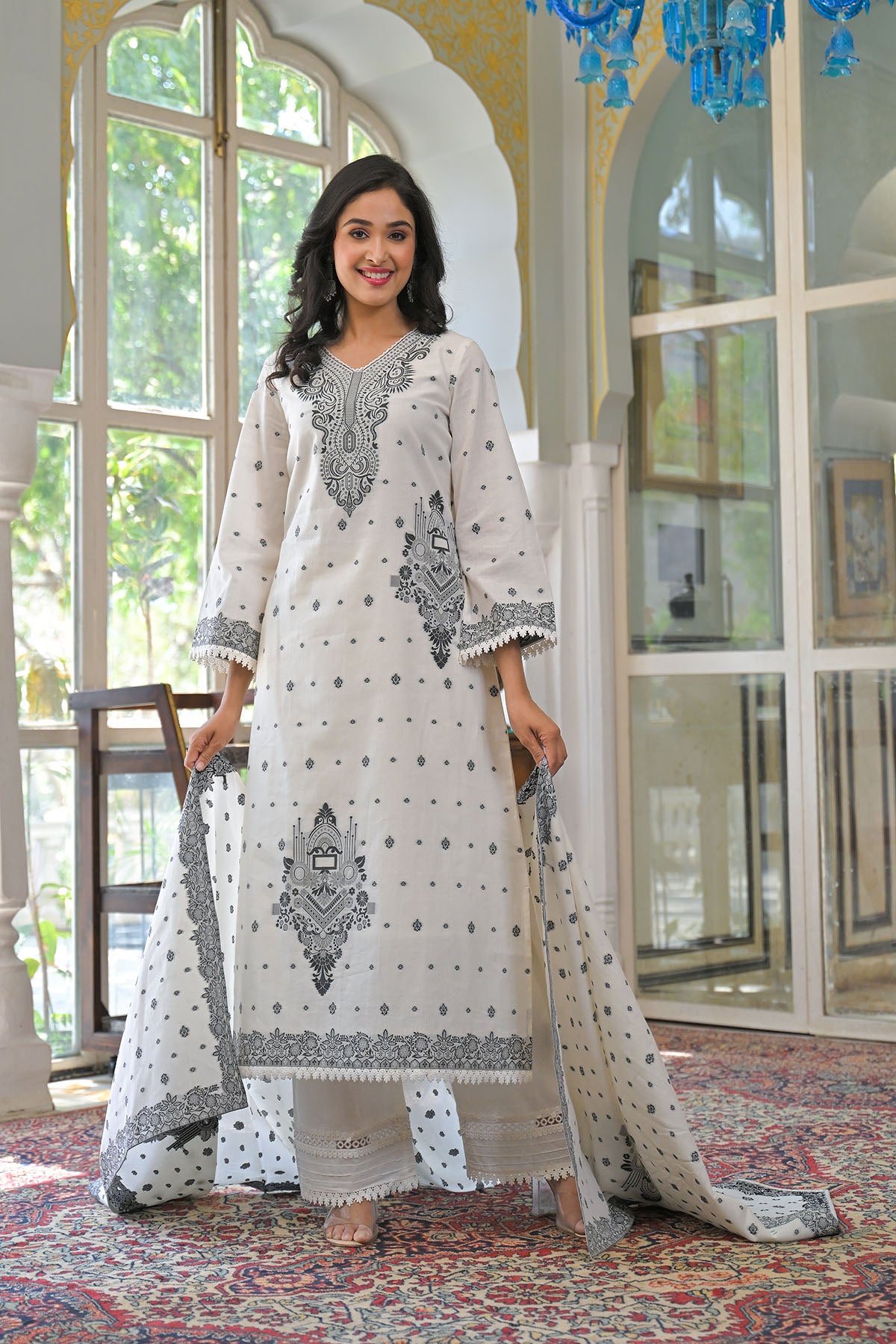 Woman in a white and gray embroidered traditional outfit standing in a room with large windows.