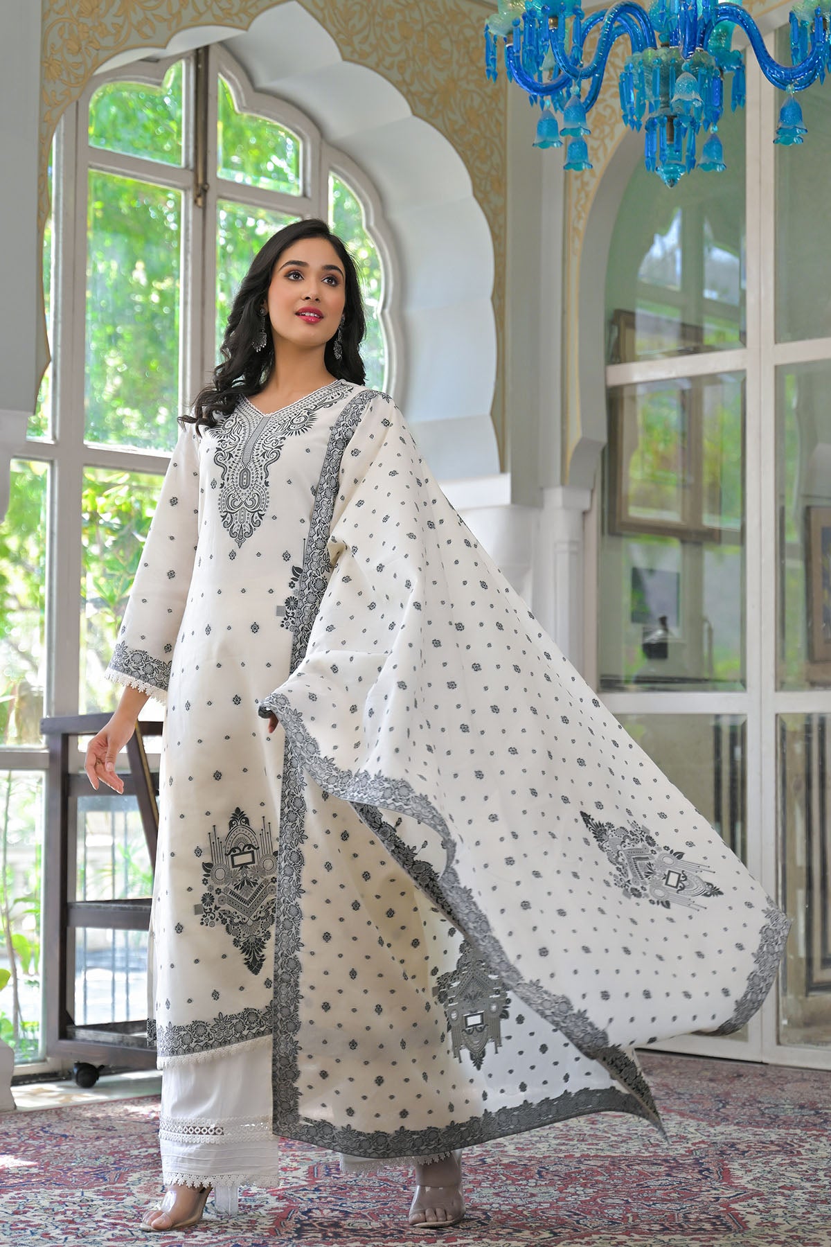 Woman in a white and gray traditional outfit standing in a room with large windows and a chandelier.