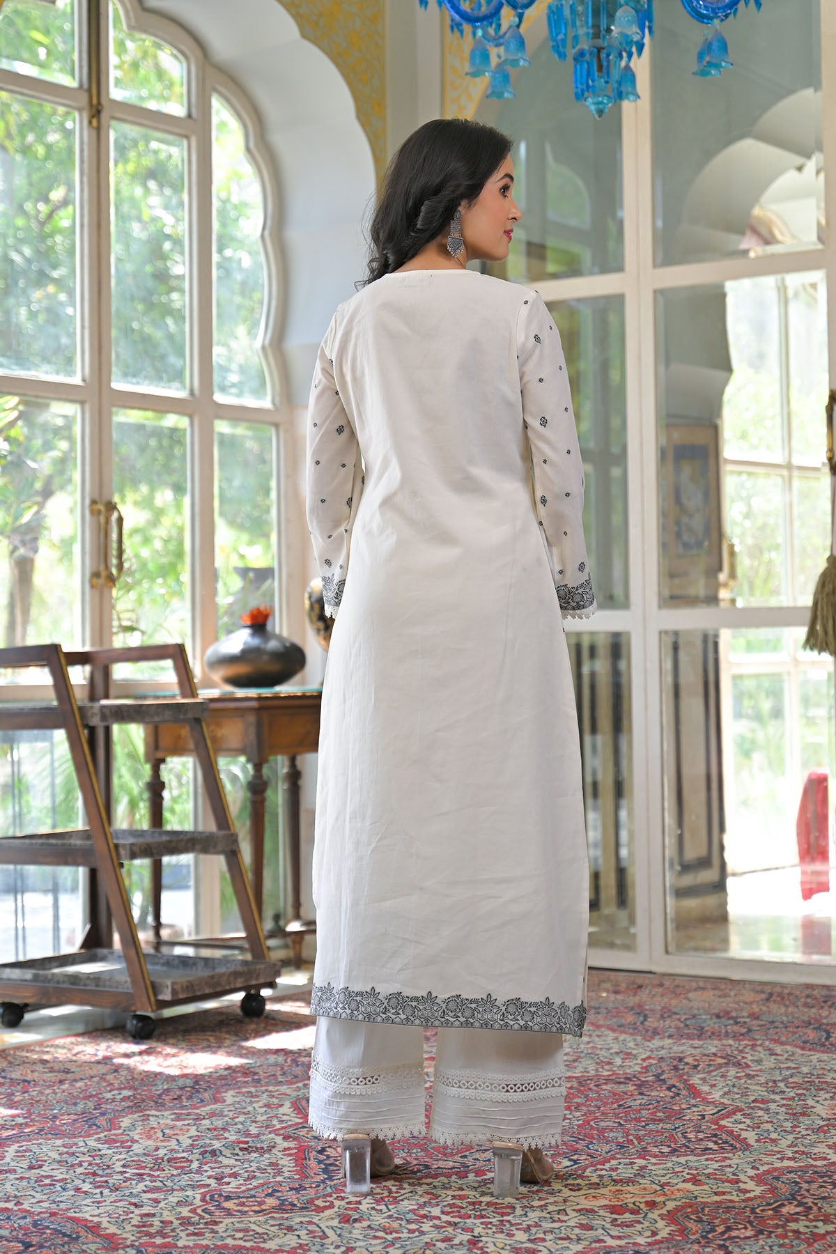 Woman in a white kurta with lace details standing in a room with large windows.