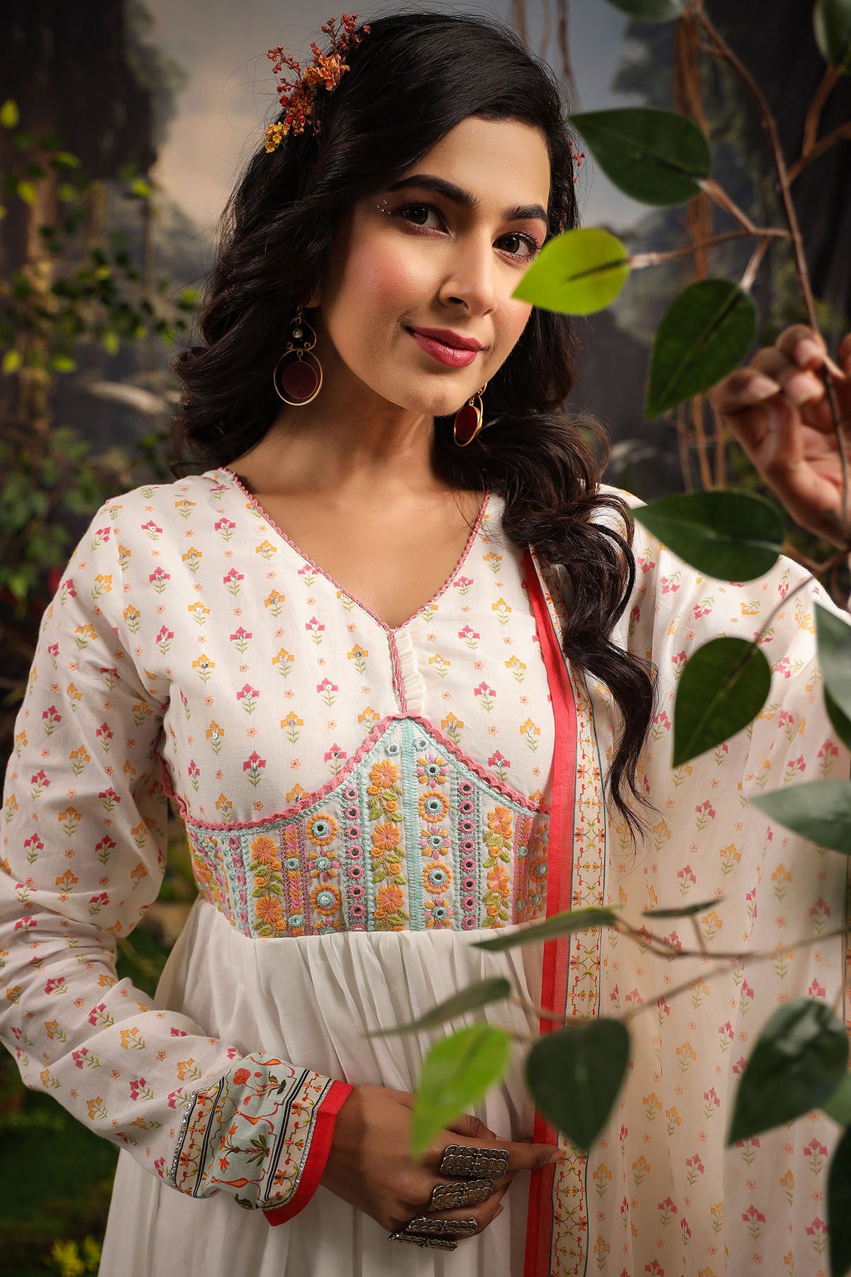 Woman in a traditional outfit with floral patterns standing among green leaves.
