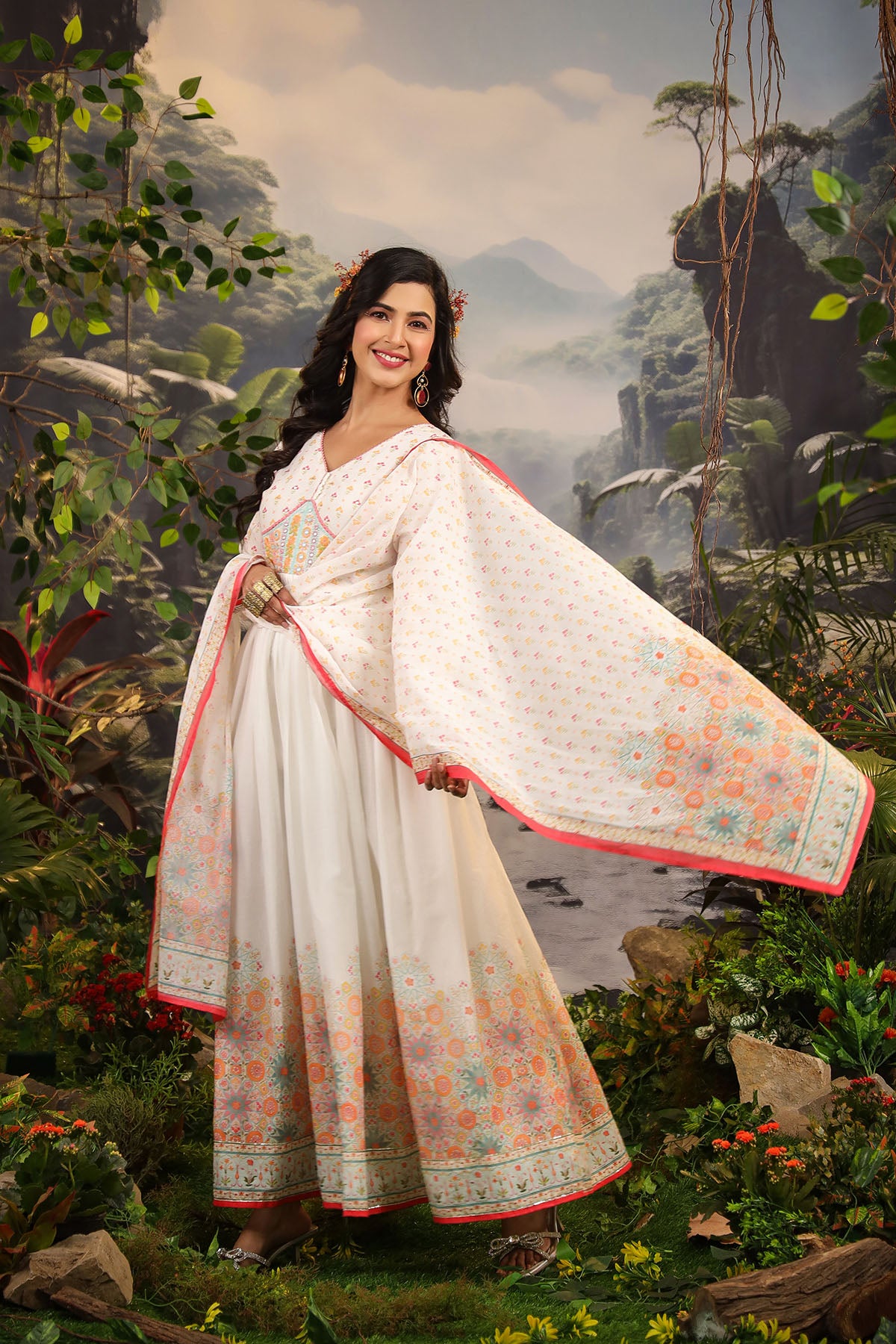 Woman in a white saree with floral patterns standing in a natural setting.