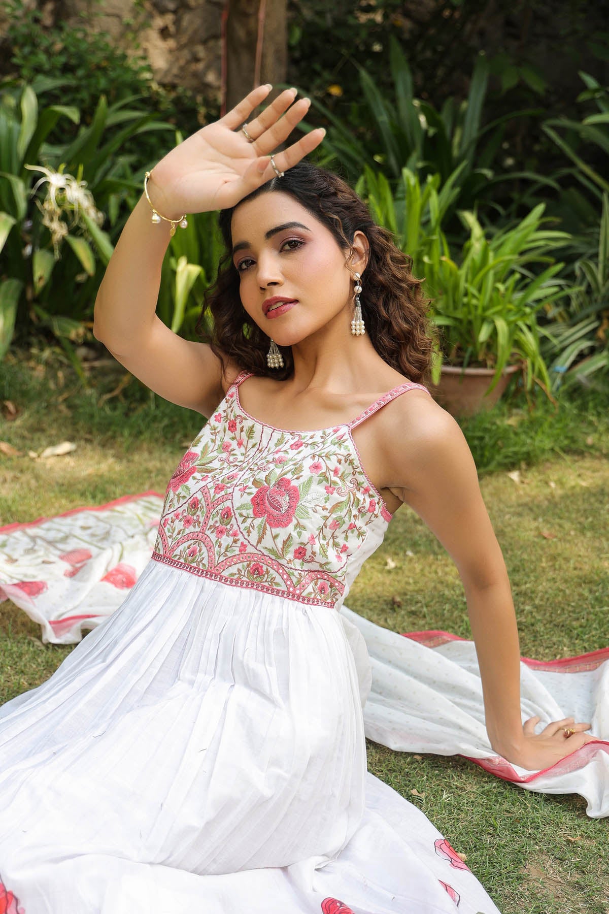 Woman in a white and pink traditional outfit sitting on grass with plants in the background