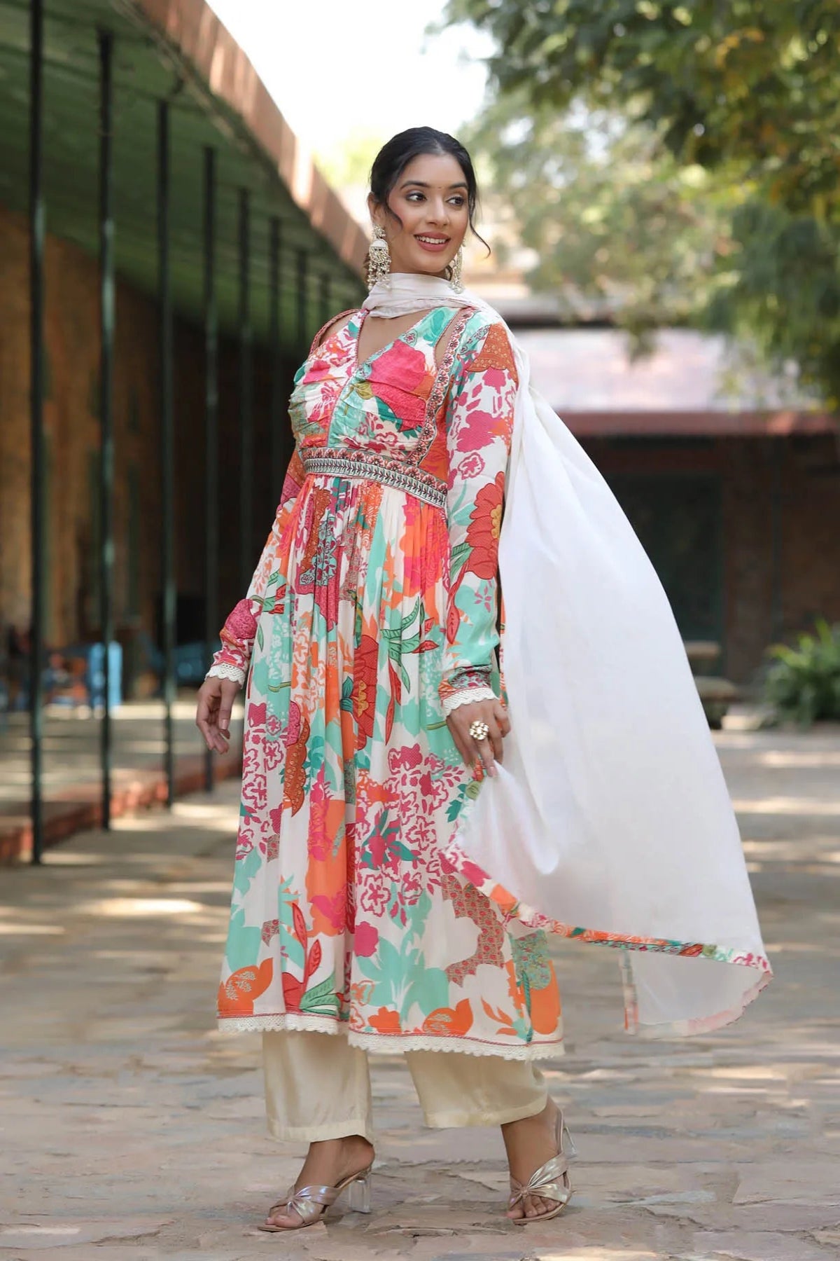 Woman in a colorful floral dress with a white dupatta standing outdoors.