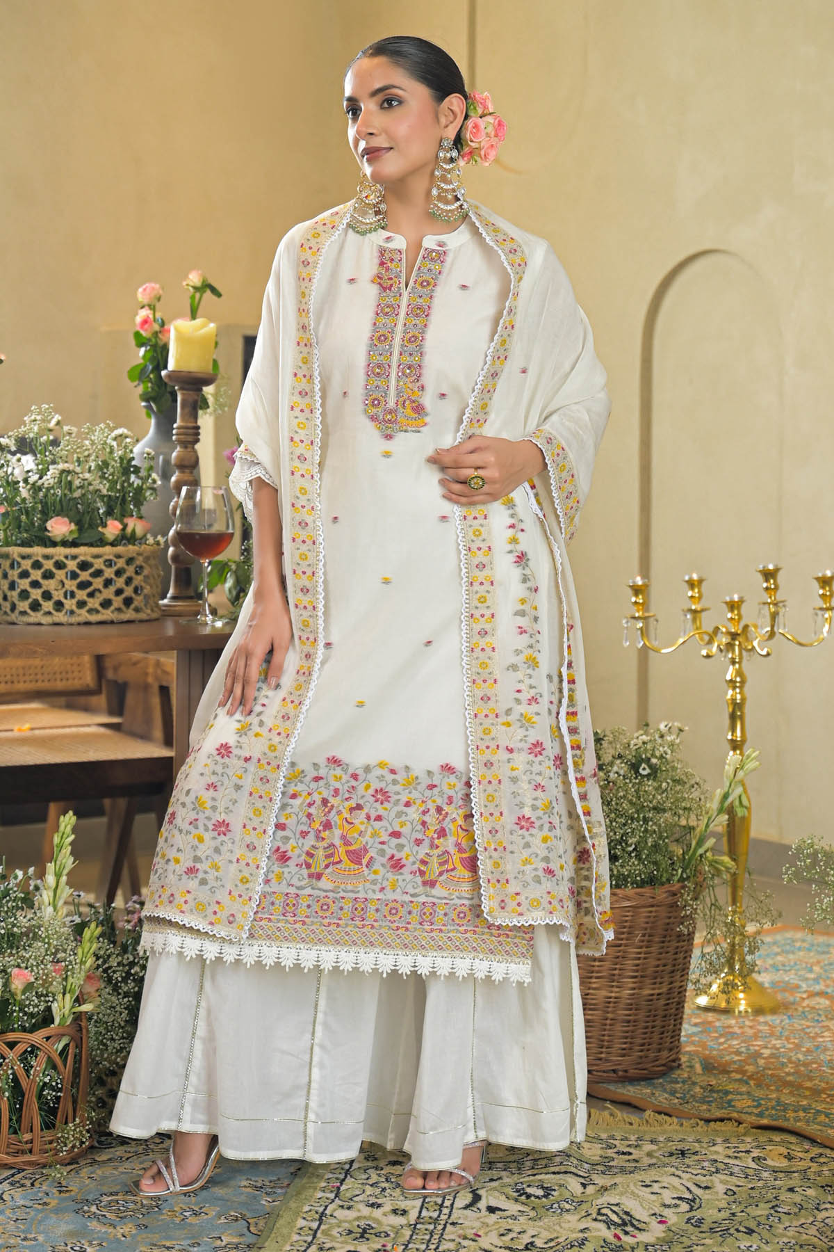 Woman in a traditional white embroidered outfit standing in an elegant room with floral decorations.