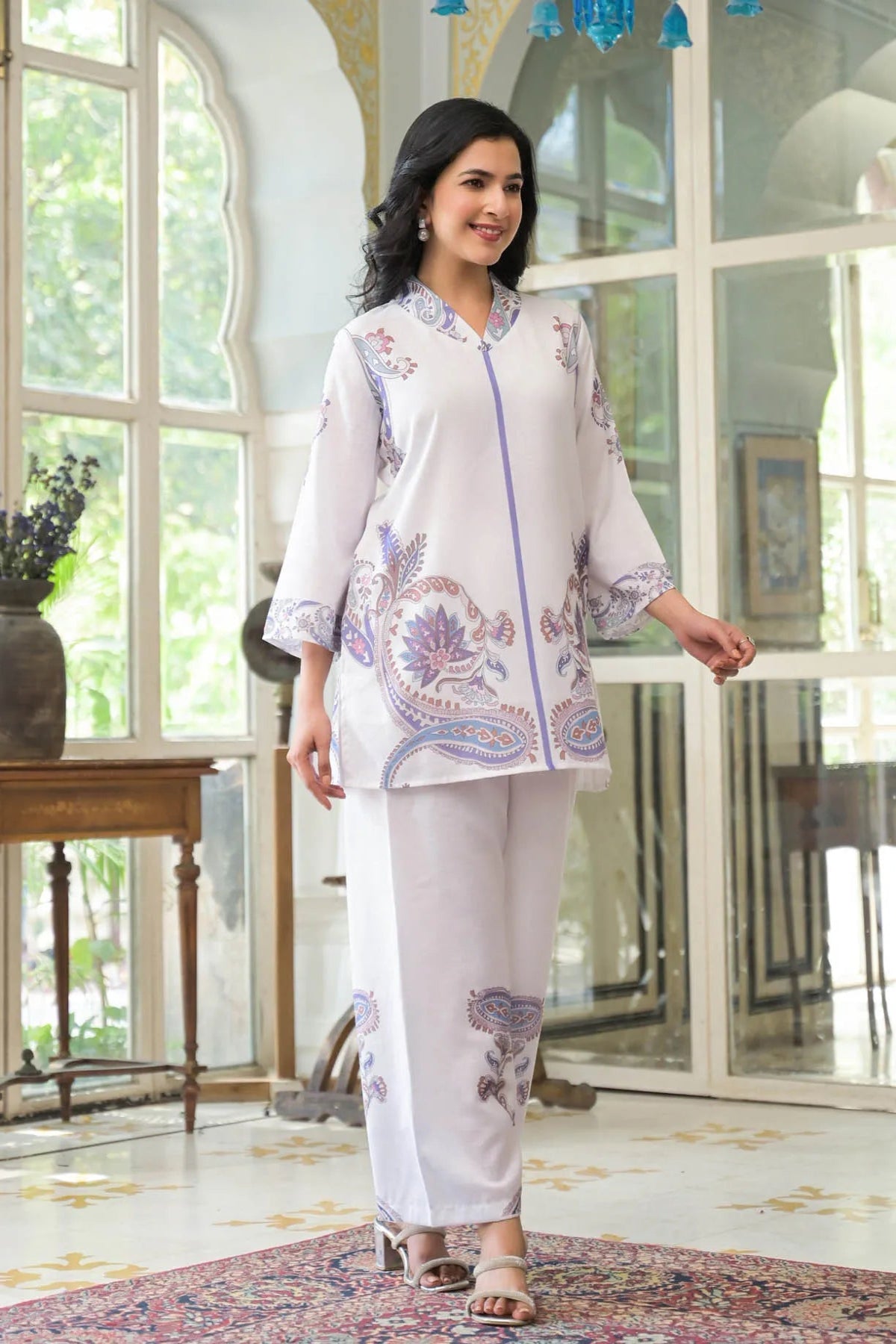 Woman wearing a white outfit with floral patterns in a room with large windows.